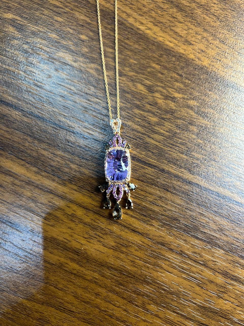 Presenta una gran amatista morada facetada en el centro, rodeada de intrincados detalles. El colgante está adornado con zafiros rosas más pequeños, diamantes y otros en tonos complementarios, que añaden profundidad y elegancia. De la parte inferior