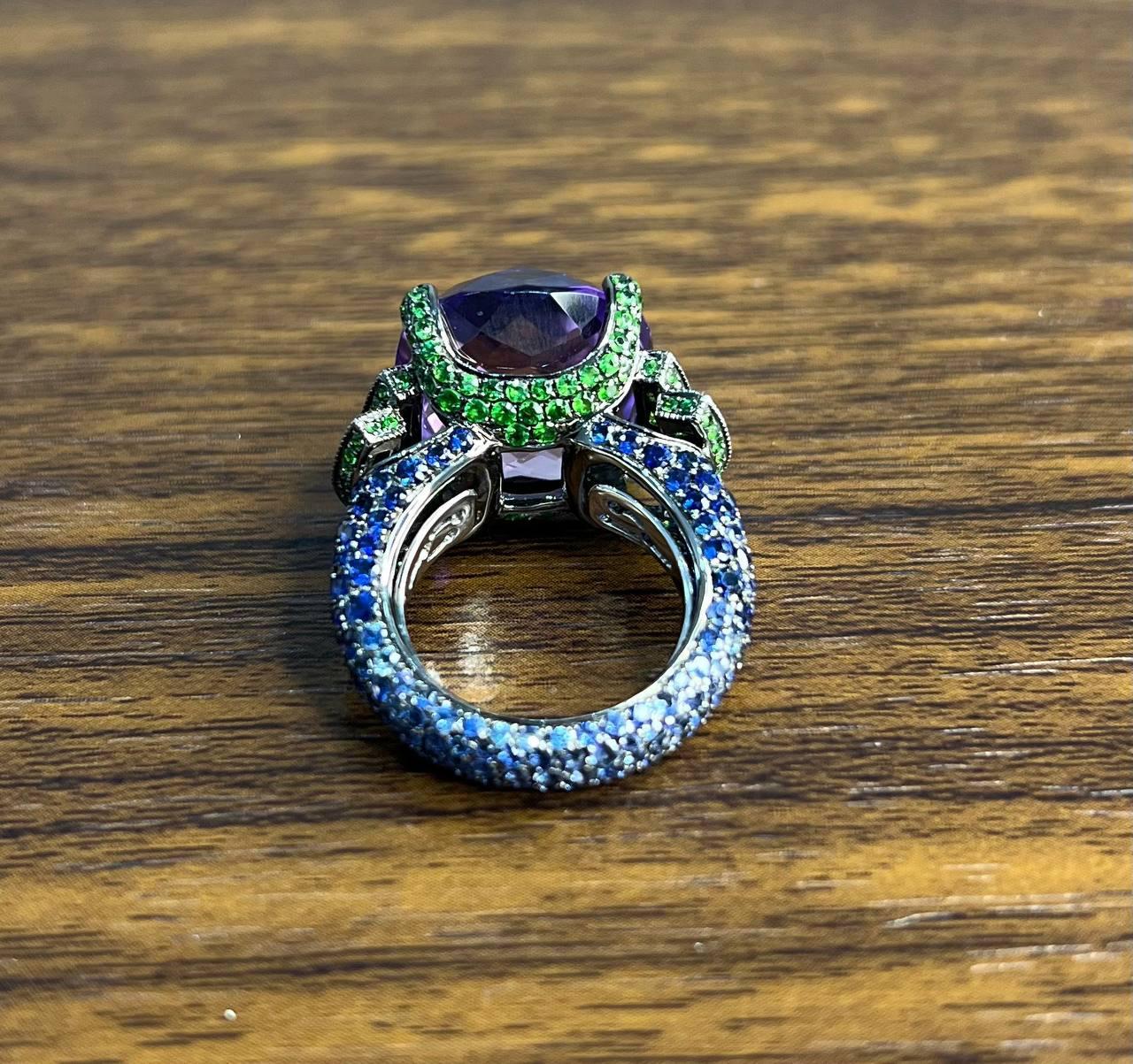 Questo squisito anello presenta una grande ametista viola sfaccettata come centro, incastonata in una montatura dettagliata. La fascia è ornata da piccoli smeraldi e zaffiri blu e verdi che aggiungono colore e contrasto. L'intricato design mette in