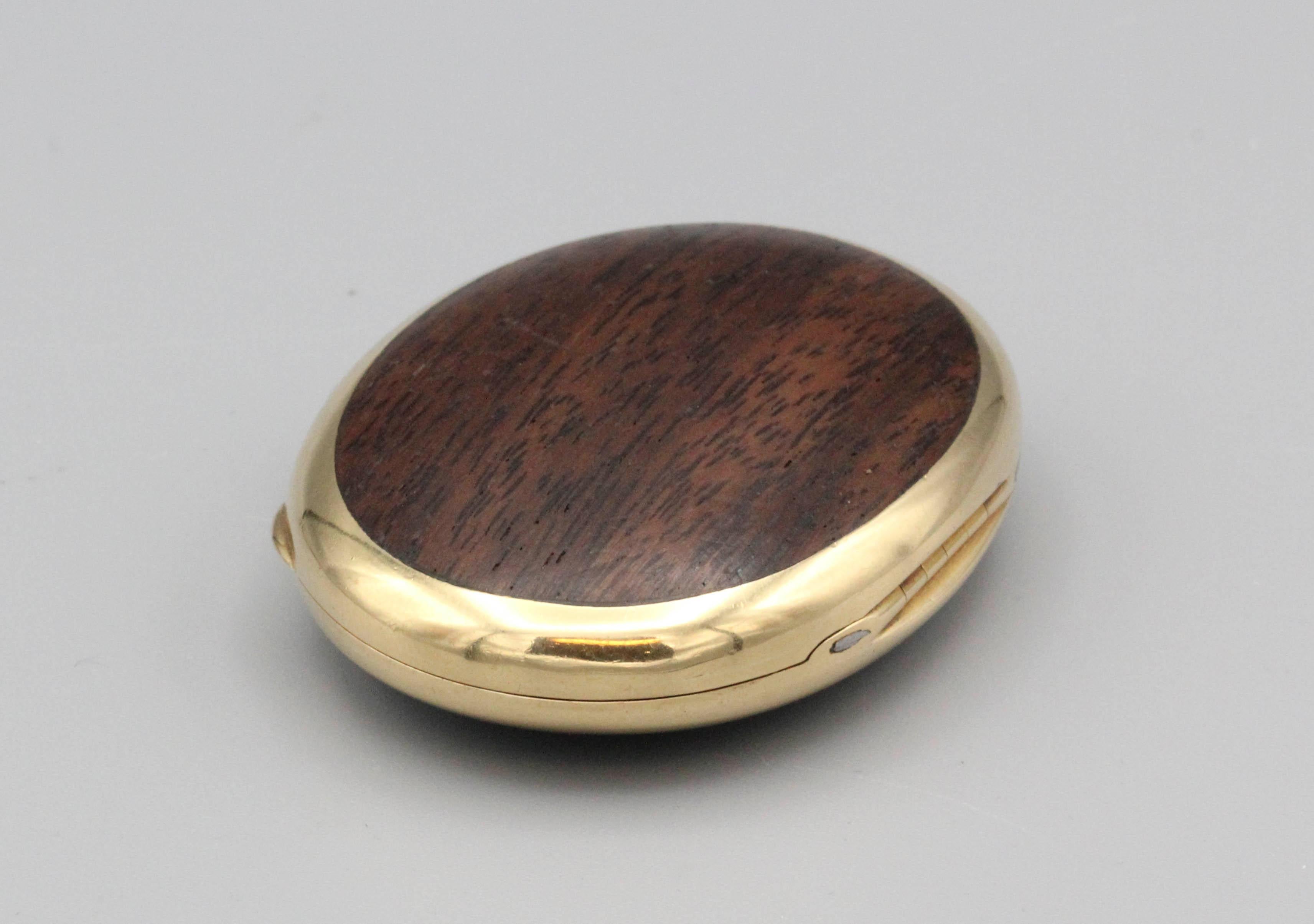 Pastillero ovalado de madera noble y oro de 18 quilates, de Cusi, hacia la década de 1980.  Fabricada en oro amarillo de 18 quilates y con incrustaciones de madera dura pulida. El pastillero se abre mediante un pulsador y pesa unos 43 gramos; mide