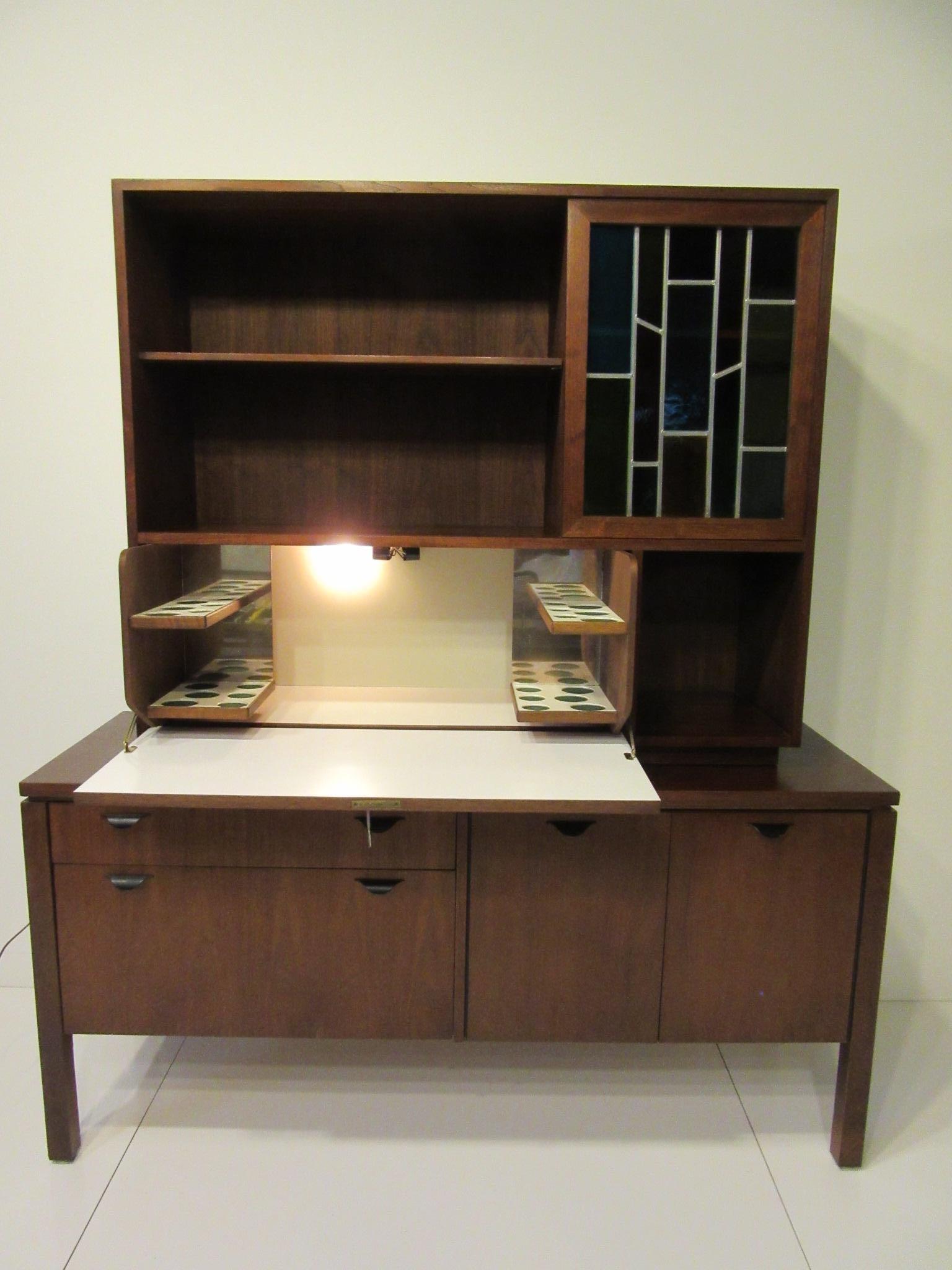 A two-piece custom bar cabinet with pull down door revealing a light up space in white laminate for glasses and bottles having the original key for the lock. The upper cabinet has two adjustable shelves, storage and a sliding stain glass door, the