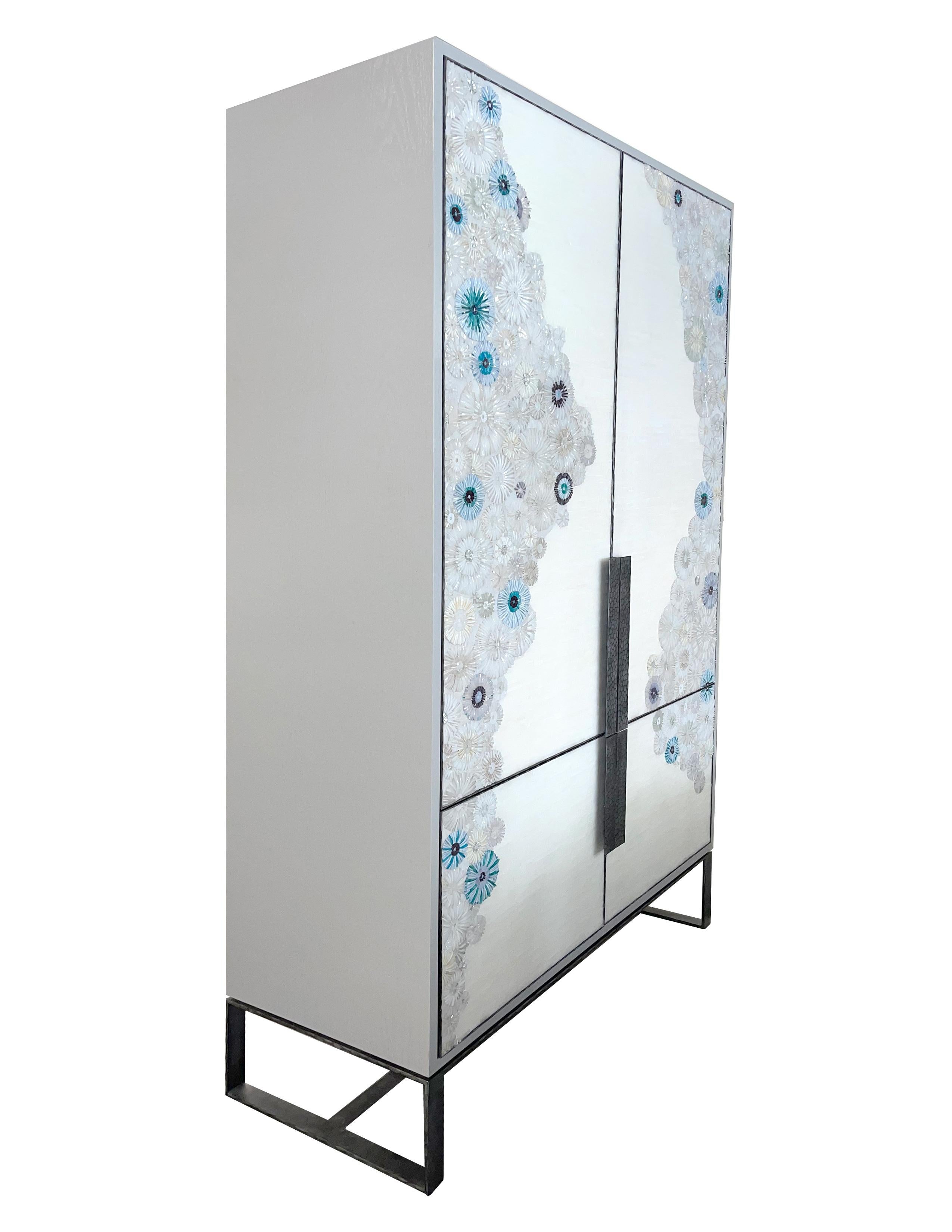 The custom Blossom Armoire by Ercole Home, is a 2-door armoire framed in hand hammered pewter metal as well as the handles. The doors are adorned in our hand cut mosaic Blossom Pattern. This mosaic consists of mixtures of whites, silvers, and wispy