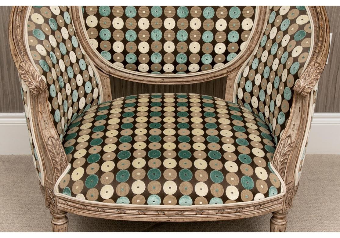 Large oval form Bergere in natural tone wood with carved Louis XVI details of acanthus leaves on the arms, floral crest and fluted legs. Upholstered in complimentary contemporary cut velveteen with raised teal, white, and tan circles on brown with