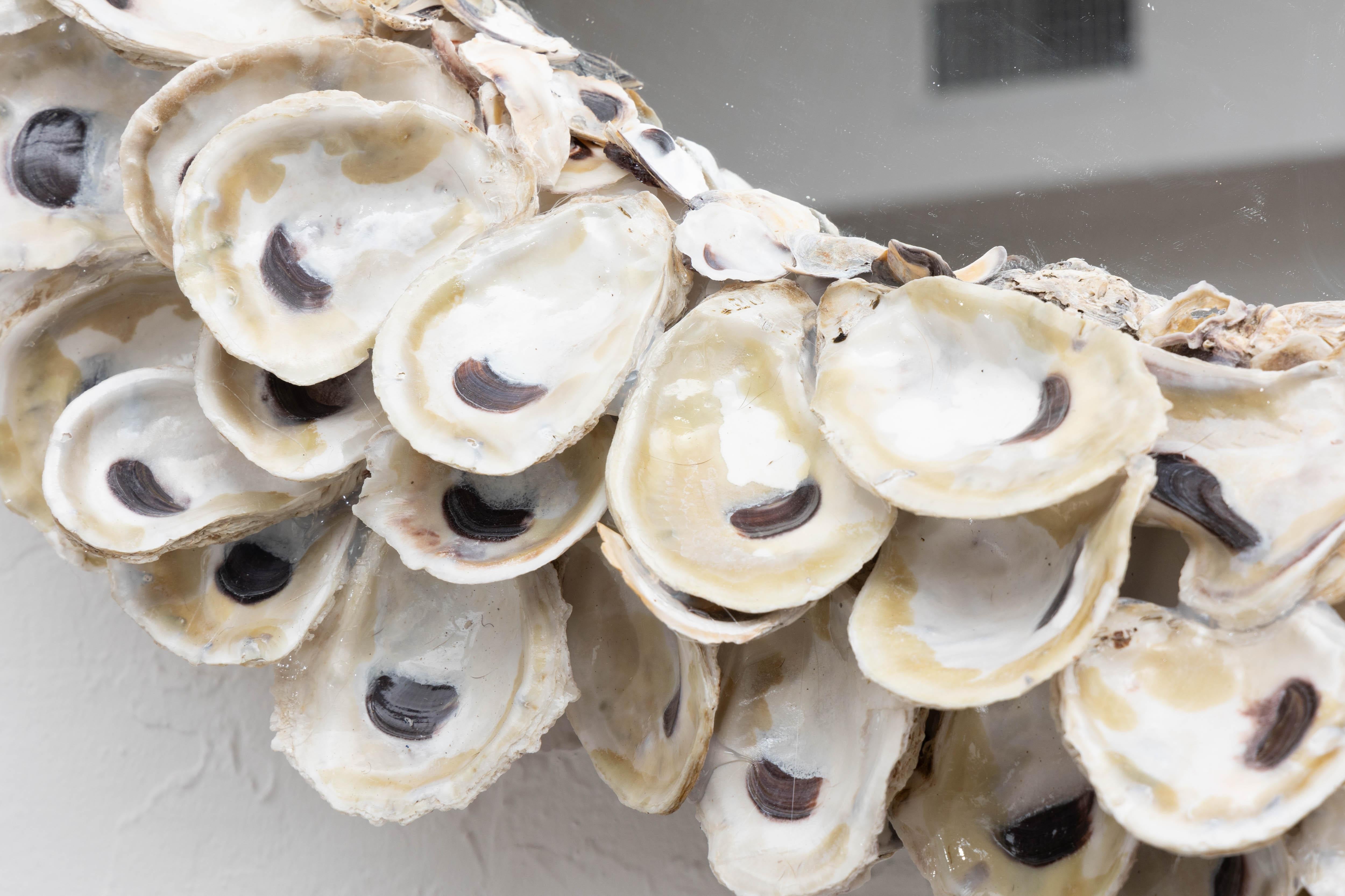 Il s'agit d'une superbe création personnalisée en forme de coquille d'huître. La plaque de miroir circulaire est entourée d'un cadre conforme constitué d'une symphonie de coquilles d'huîtres organiques.