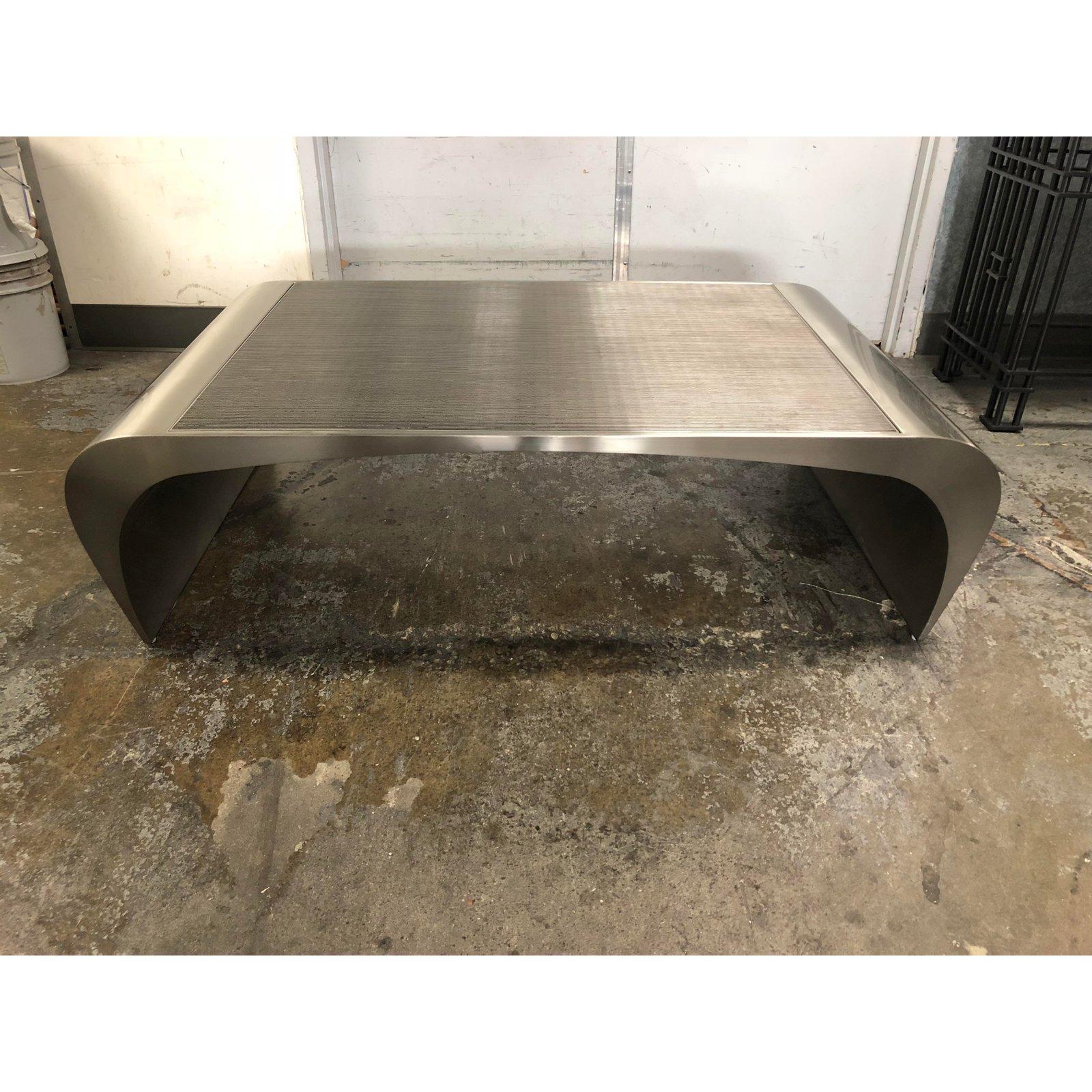 A curved, brushed steel coffee table with a metal grate top. Durable and industrial, this piece sports minor blemishes on the metal finish. Like a jet engine that just landed in your living room, this piece boasts curved and flared ends, streamlined