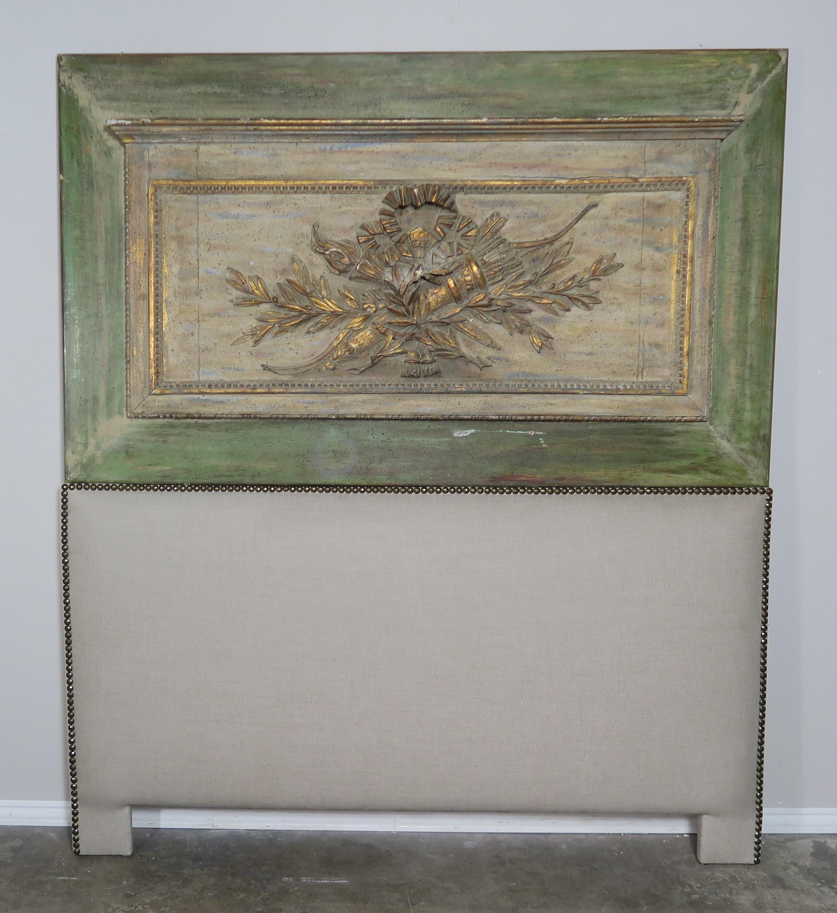 Custom queen size headboard made with a 19th century painted and parcel gilt panel depicting torches, laurel leaves and ribbon. Beautiful tiny carved beading frames the scene. The panel has been upholstered into a queen size headboard using Belgium