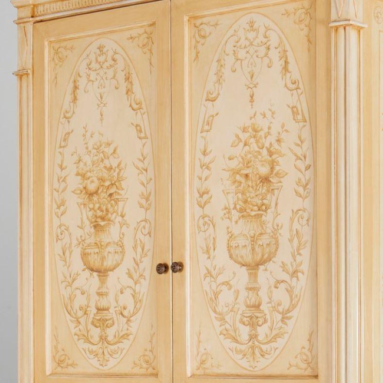 20th c., A beautiful custom made Louis XVI style armoire, hand painted with a flowering urn motif, in pale beiges on a light cream background. The upper cabinet interior has been fitted with two shelves and has space for electronics. This section