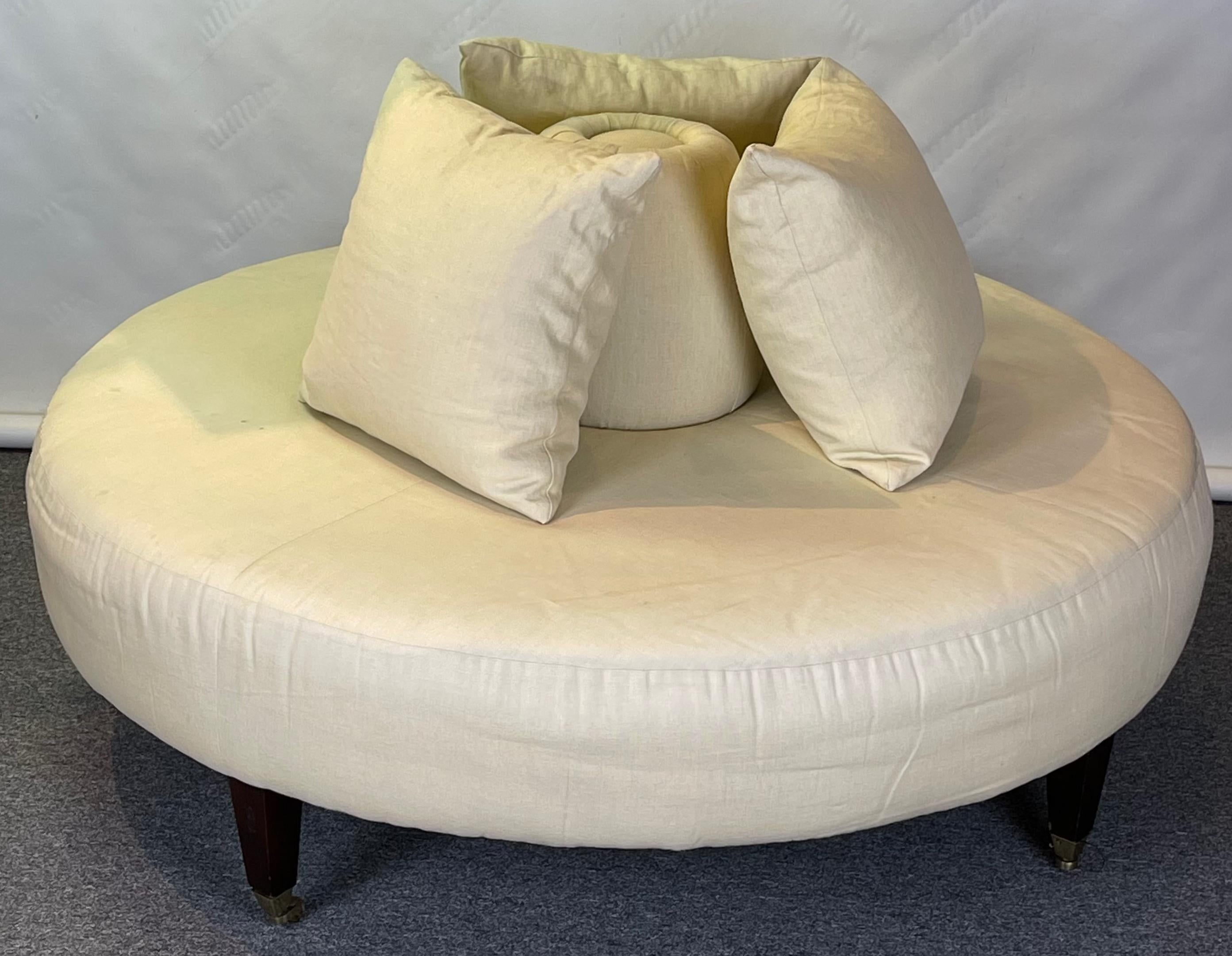 A custom upholstered circular confidante or round bench upholstered in off white linen resting on four square tapering legs with brass cup casters.