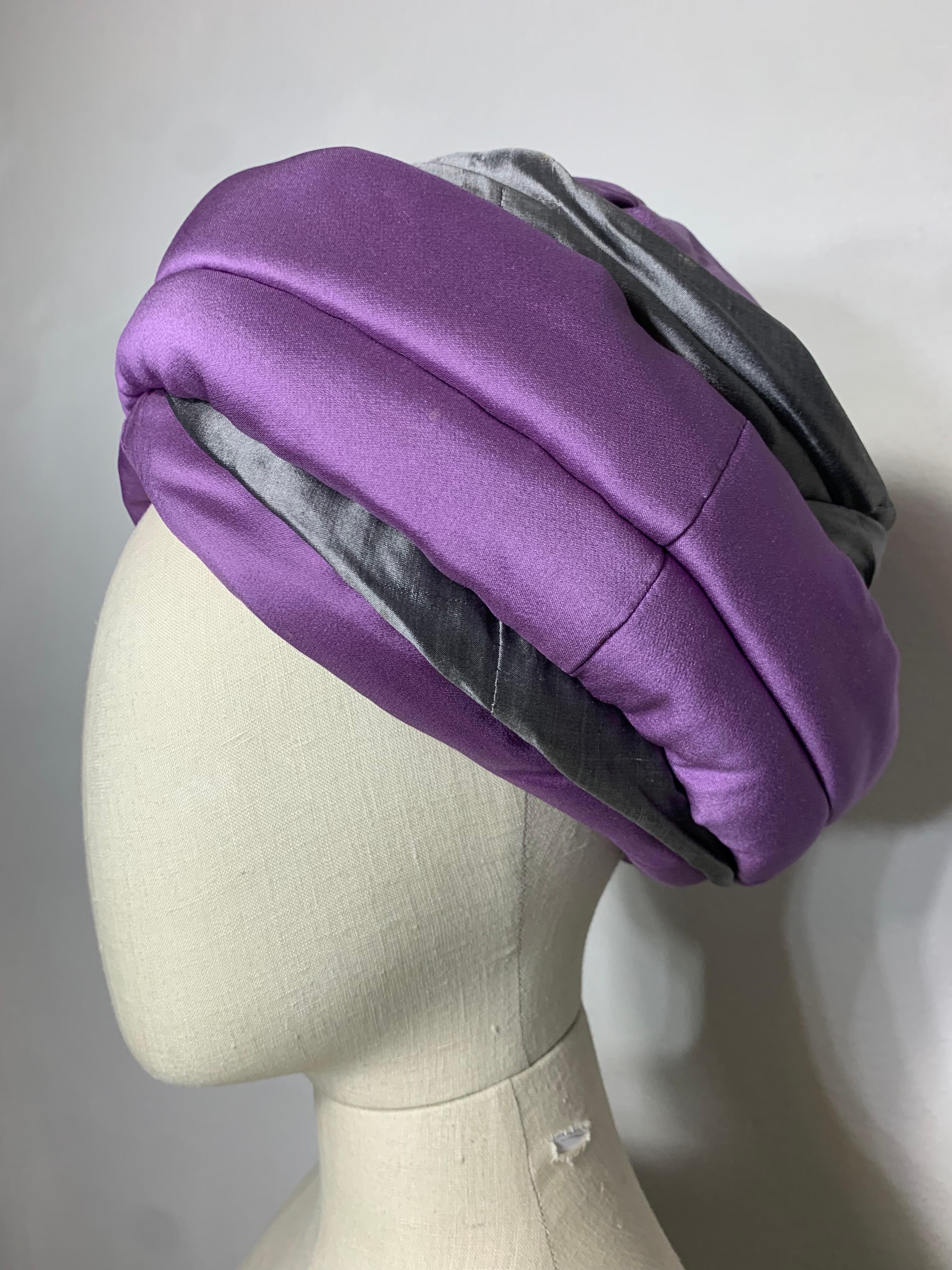 Suzanne Couture Millinery su misura, viola e grigio, con ciuffo e drappeggio, turbante con spilla per cappello: corona leggermente spiovente. Strutturato su una base in buckram per conferirgli altezza e stile. La pelle di squalo in seta viola e