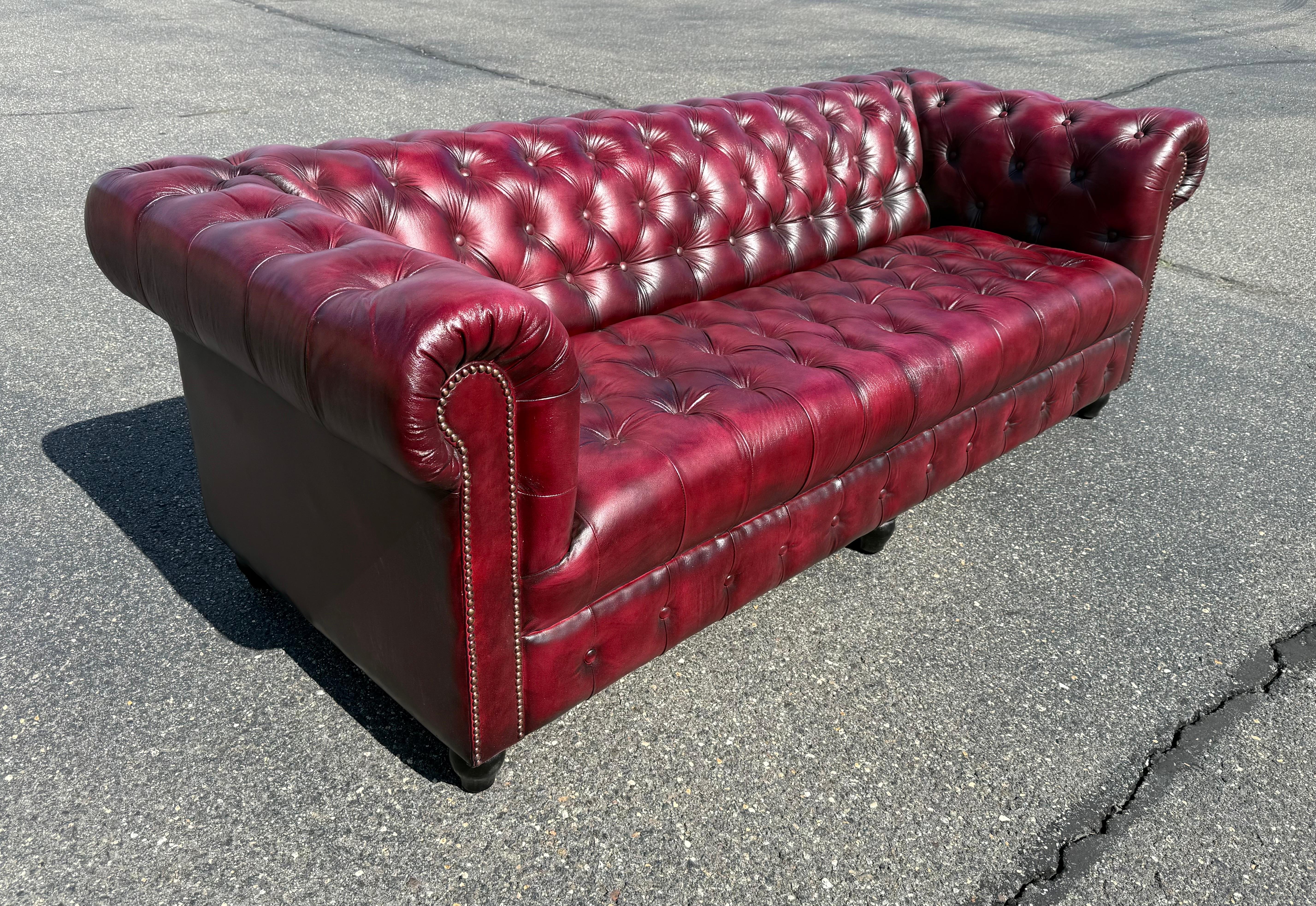 This handcrafted English Chesterfield sofa in oxblood genuine leather is a stunning example of timeless design and traditional craftsmanship. Upholstered in a rich burgundy leather, the sofa features a dramatic tufted bench seat, classic deep