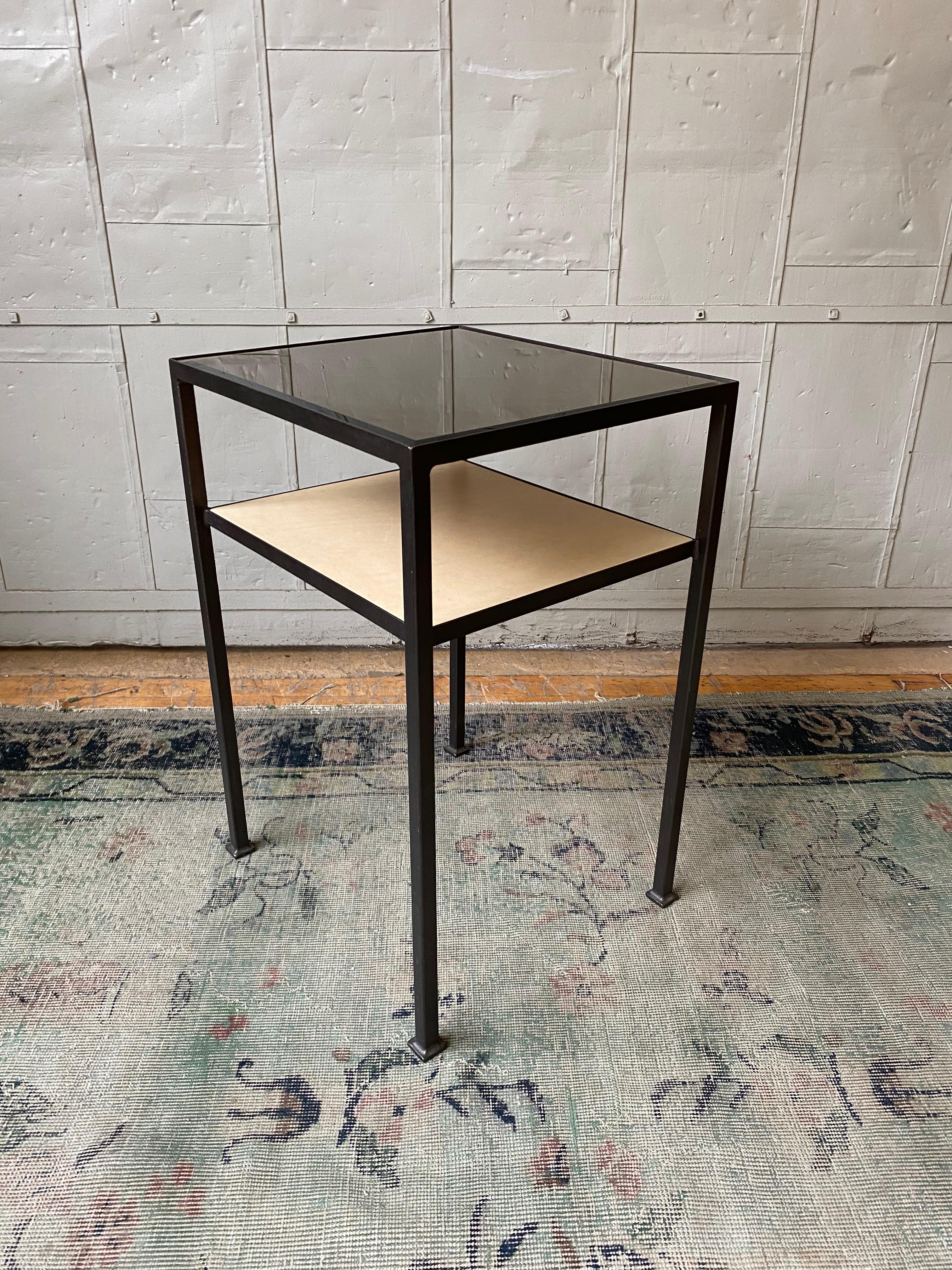 Cette table de nuit ou table d'appoint bien proportionnée présente un cadre en fer avec une finition bronze appliquée à la main, un plateau en verre transparent et une étagère inférieure recouverte de cuir. Il s'agit d'un échantillon de sol de notre
