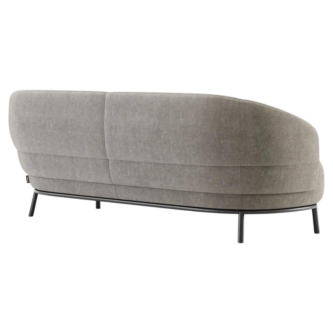 This sofa is a rounded piece supported by four steel legs and tubing. Characterized by soft curves and balanced proportions, the backrest extends to the sides creating subtle and elegant armrests. The highlight of this piece is represented by the