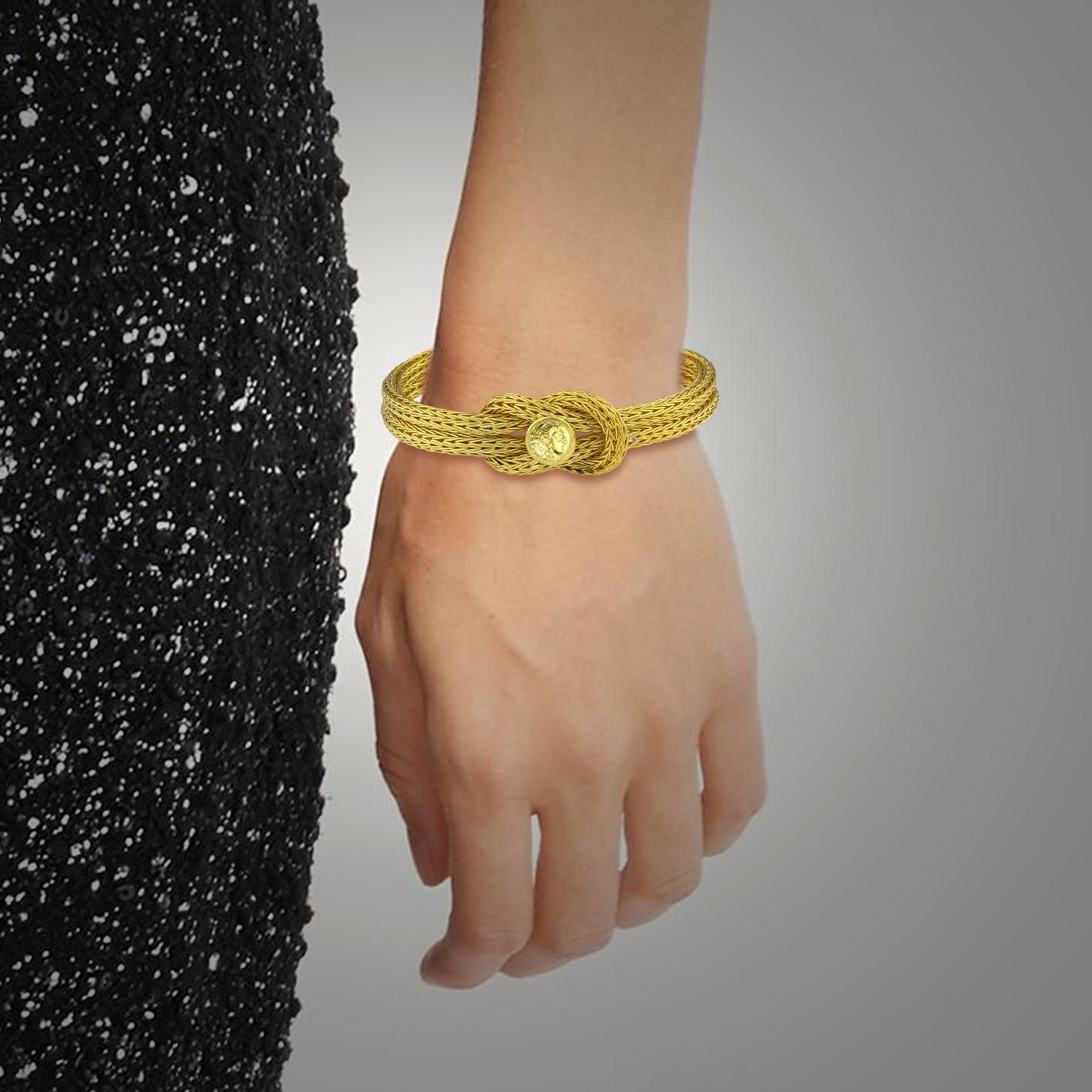 S.Georgios Designer-Armband, handgestrickt in unserer Werkstatt in Griechenland aus 18 Karat Gelbgoldfäden. Dieses flexible Armband ziert den Herkulesknoten, das Symbol der Stärke, der Heilung und des Schutzes, sowie eine Replik einer Münze von