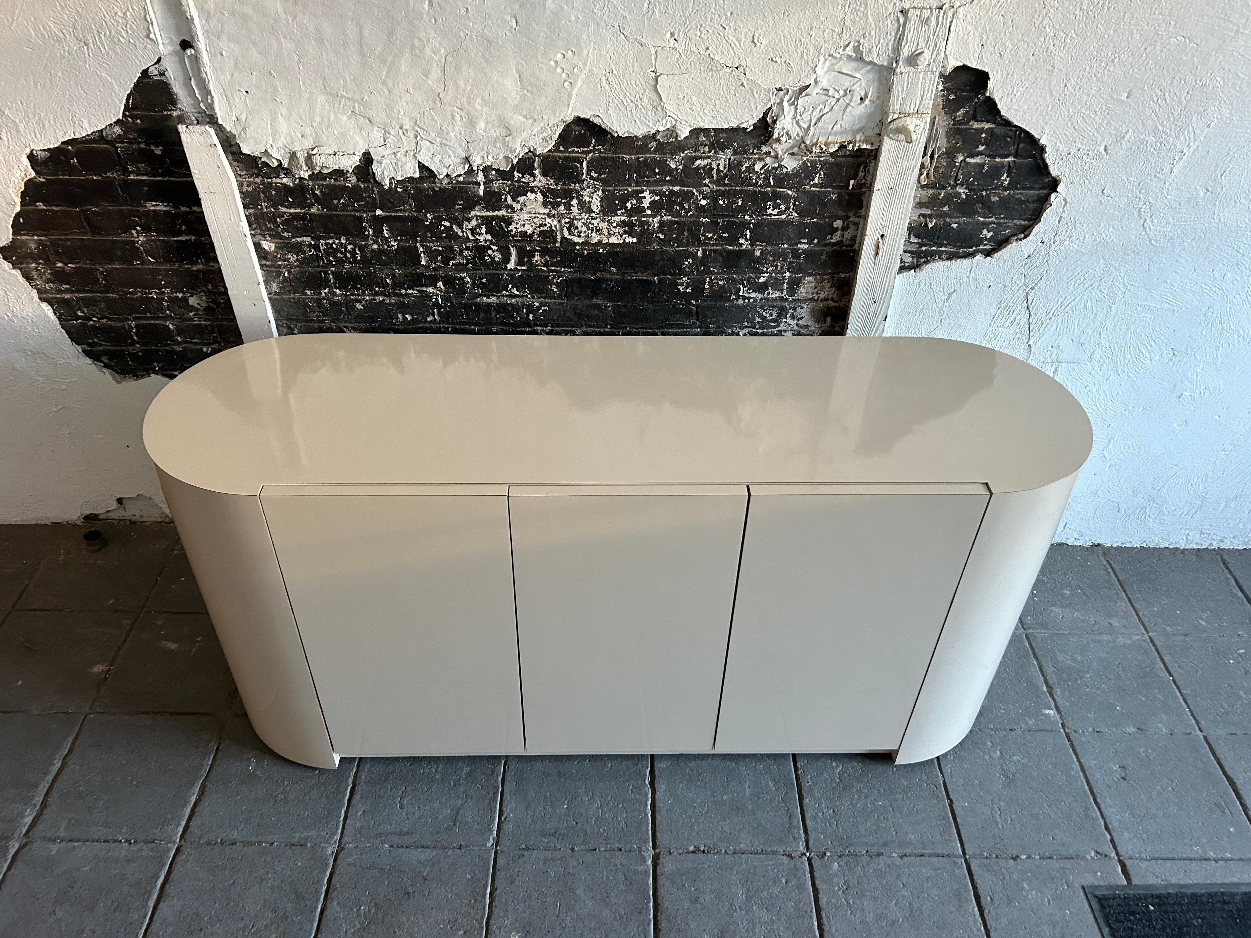 Pop art post modern 3 door beige colored Pill shaped rounded ends credenza sideboard. Very light tan beige color gloss laminate credenza custom made circa 1980 has 3 cabinet doors with catches. Behind the doors are (2) adjustable shelves. Ready for