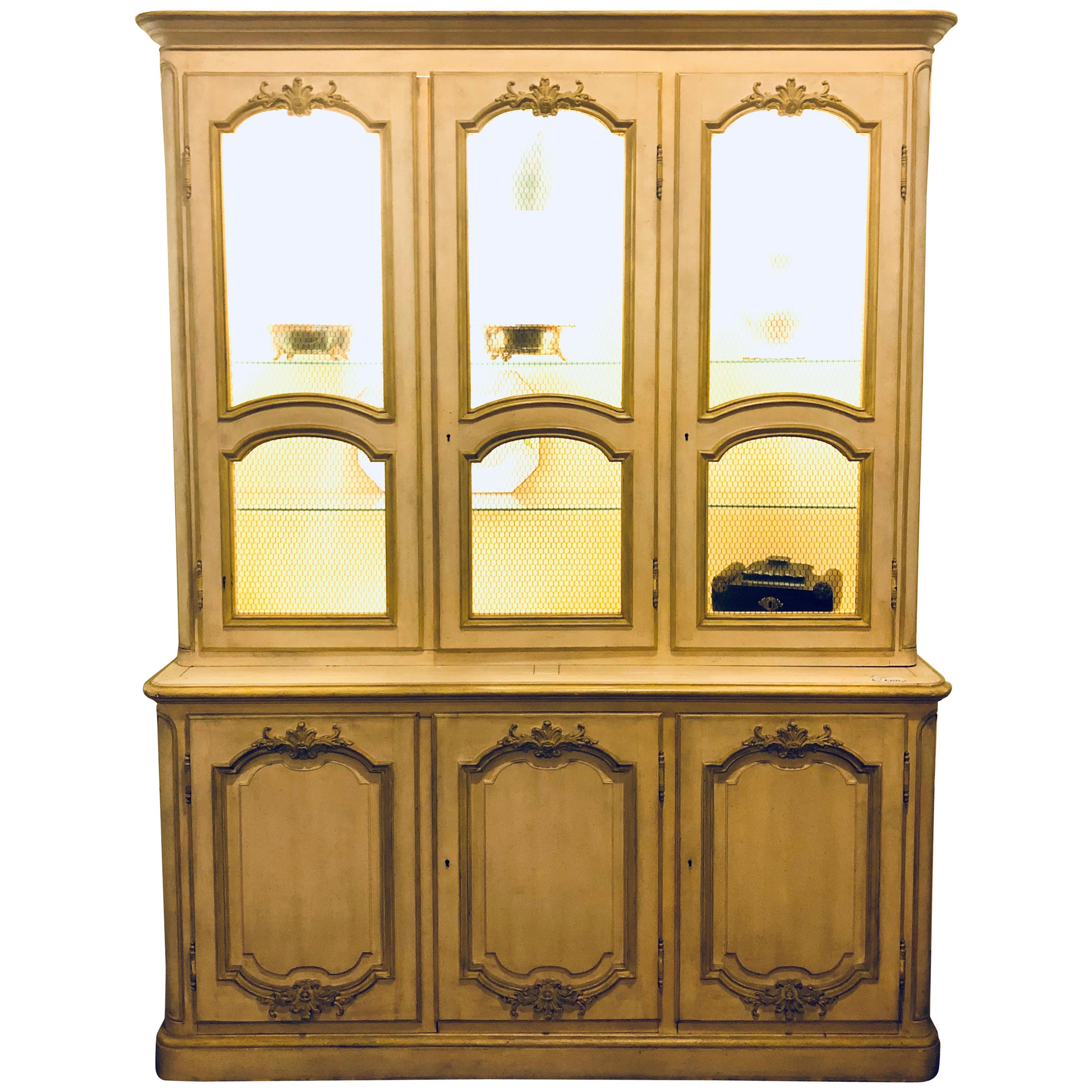 A custom quality Hollywood Regency baker cream and gilt distressed china cabinet oak lined. This fine distressed two-piece breakfront china cabinet is certain to light up any room in the home or office. On sale.