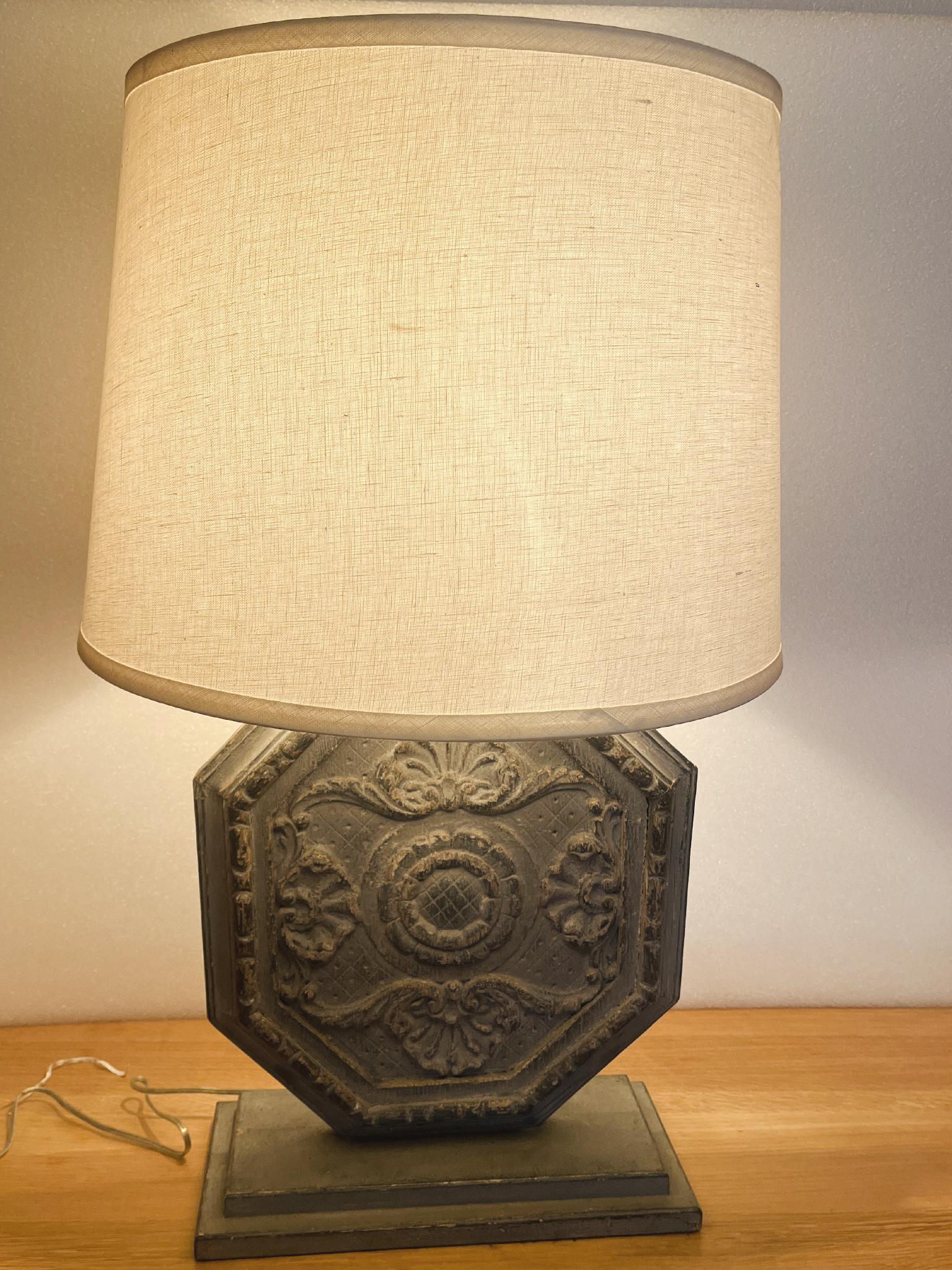 Cette charmante lampe est fabriquée sur mesure et se compose d'un bouclier hexagonal en bois du début du 20e siècle surmontant une base rectangulaire en bois. Des motifs floraux ornent la face du bouclier. La lampe est assortie d'un abat-jour en lin