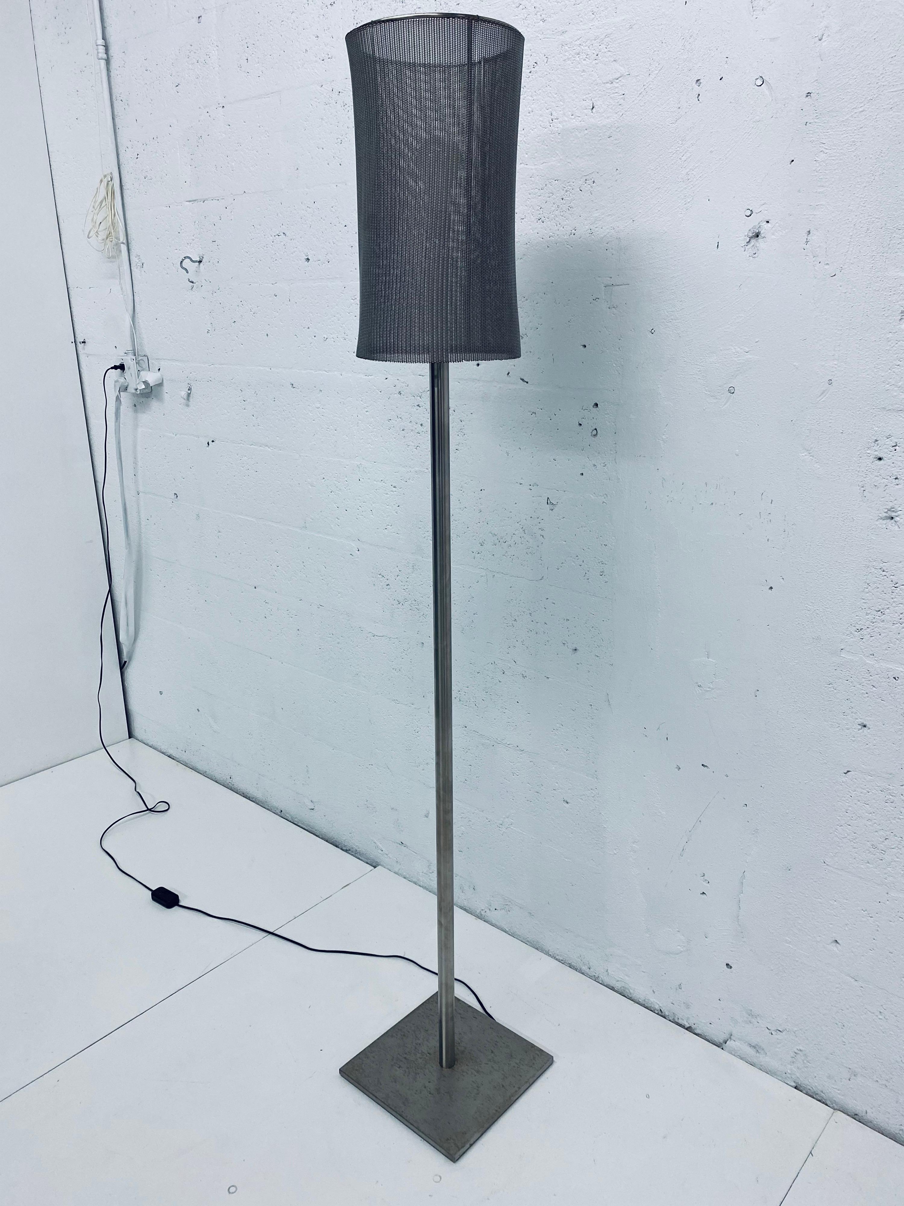 Custom floor lamp fabricated of a tubular steel rod welded to a square base and topped with a metal mesh lamp shade. The lamp shade frame is welded and not removable and the mesh is attached via metal clips. Light is dimmable. Retains label: