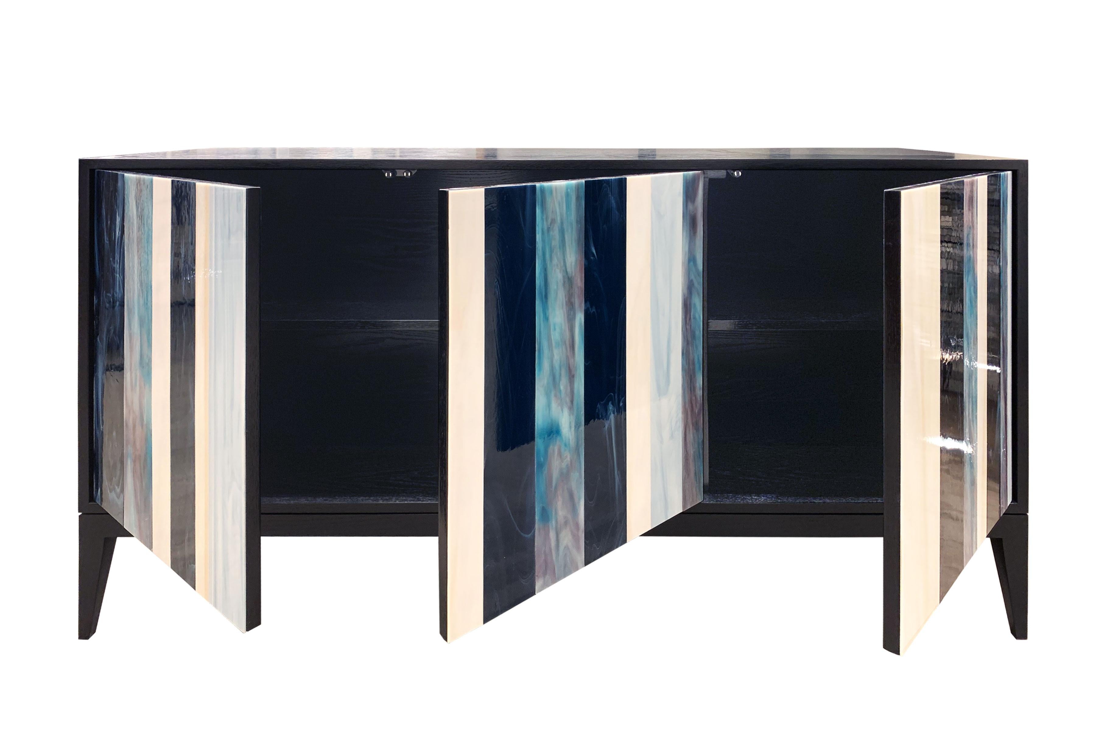 The Milano Linea buffet by Ercole home has a 3-door front, with black stain finish on oak.
Handcut glass stripes in Ivory, French Blue, Electric Blue, and Cloudy Notte decorate the surface of doors.
There is one adjustable interior shelf in each