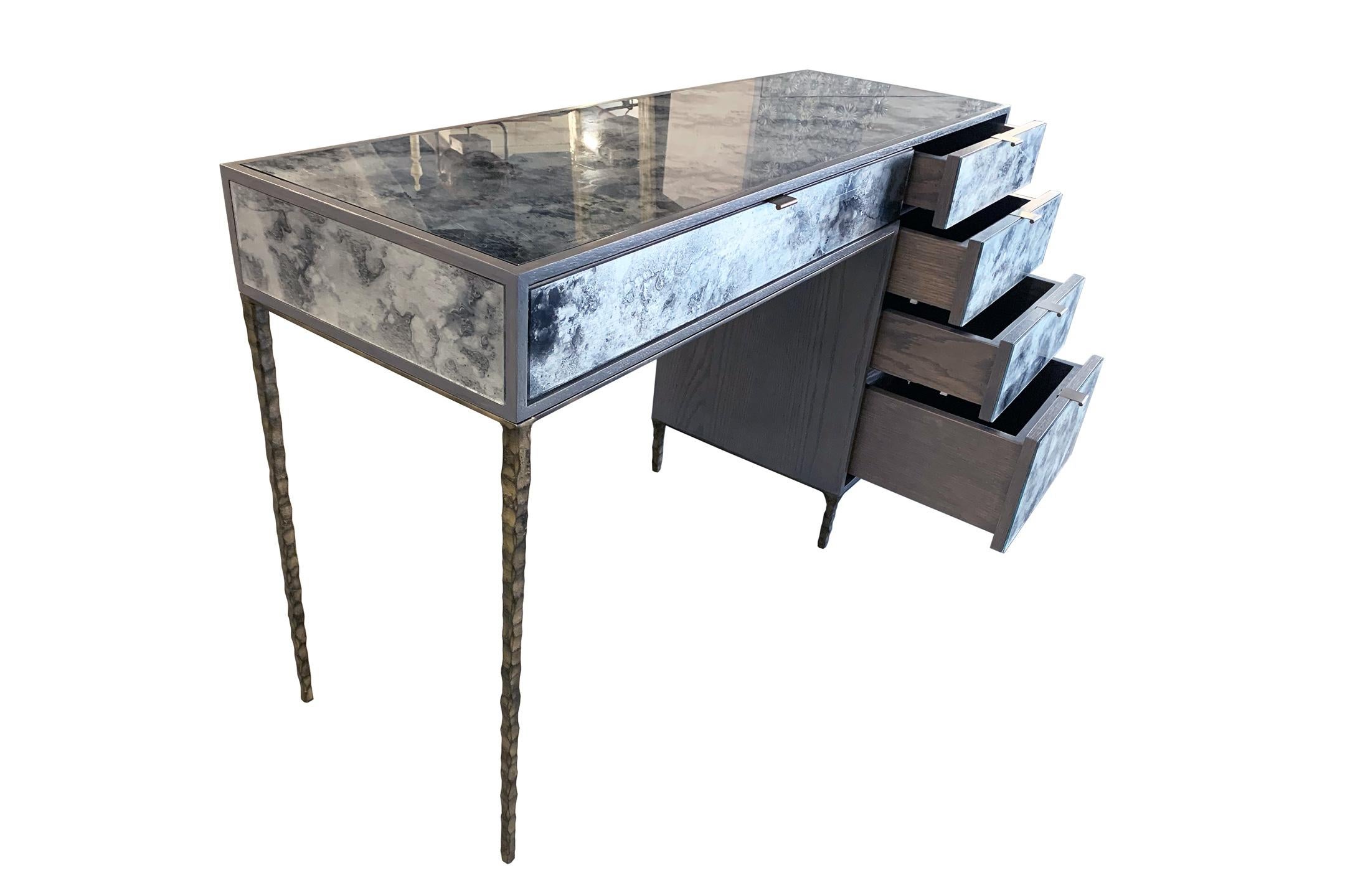 The mystic fume silver vanity by Ercole Home has a 5-drawer, with Earl gray wood finish on oak. Hand painted églomisé glass panels are inset on the surface.
There are five aluminum pulls and the hand-hammered metal base are in bronze