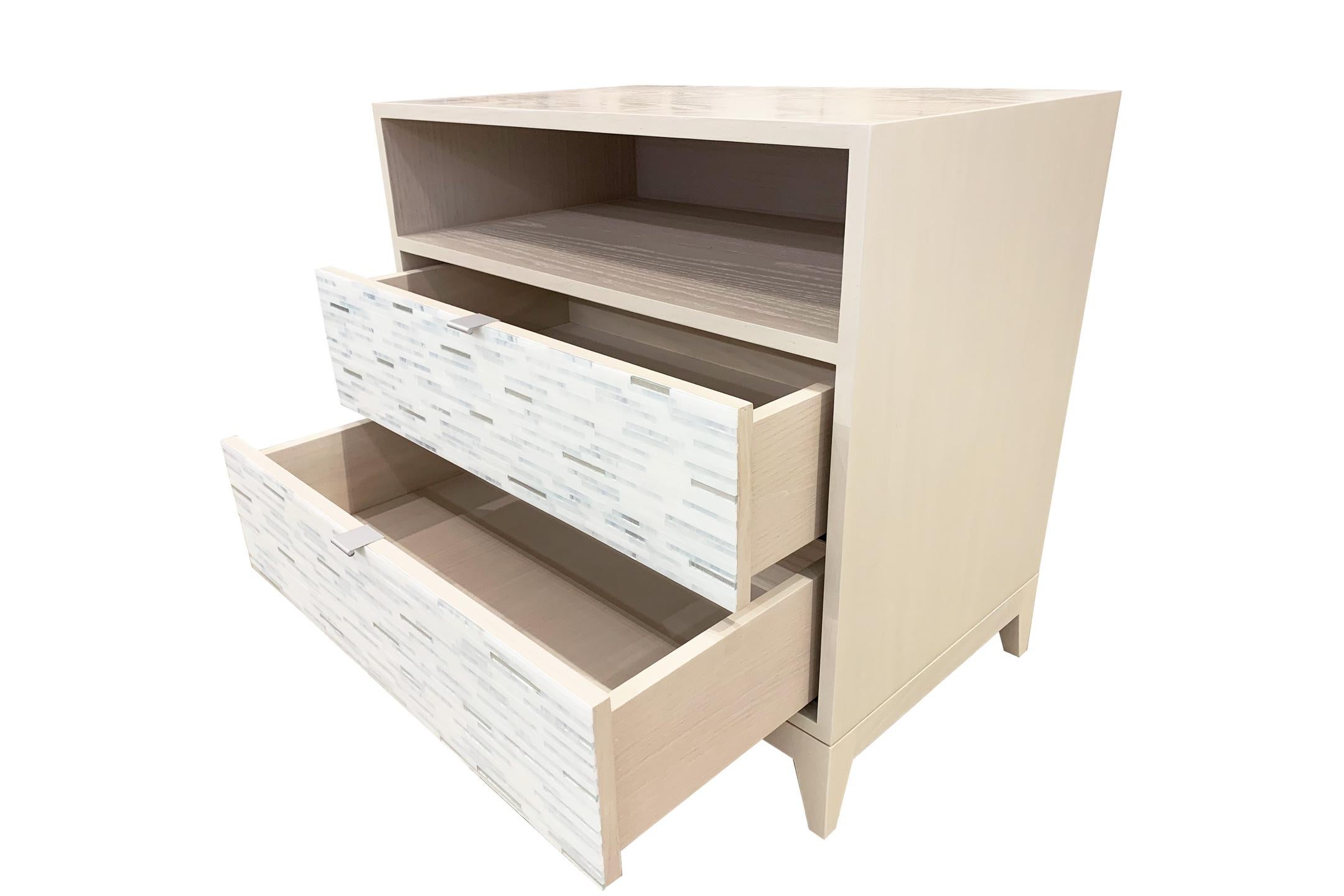 The Milano nightstand by Ercole Home has a 2-drawer front and open shelf, with washed ivory wood finish on oak. Handcut glass mosaics in icy white, wispy white, and silver decorate the surface in a horizontal regular stripe pattern. There are two