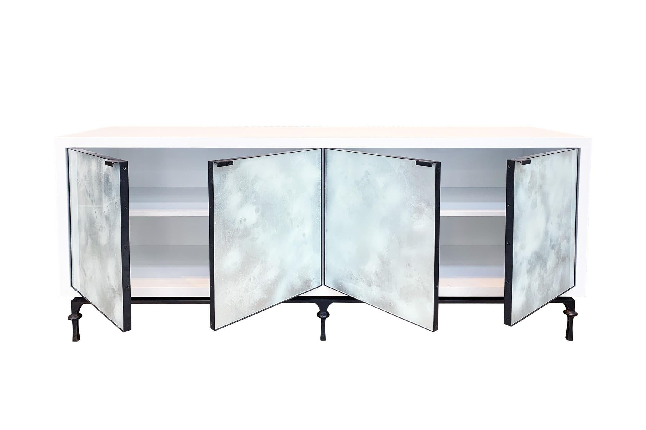 The Mystic buffet or credenza by Ercole home has a 4-door front, with painted white wood finish in matte sheen.
Hand painted glass panels in Oro Bianco are on the door surface.
The decorative hand forged Meta base in natural steel metal