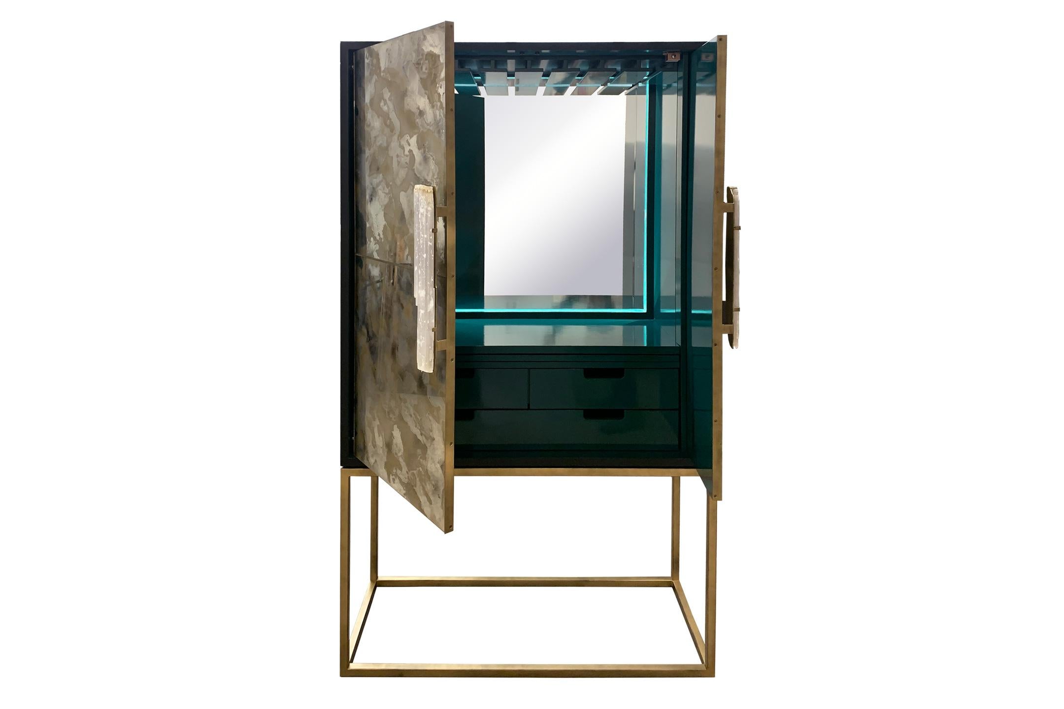 The gold dust bar cabinet by Ercole Home has a 2-door front, with matte black exterior and green lacquer interior wood finish.
Hand painted Églomisé mirror in gold dust decorates the surface with Selenite handles.
Hand forged metal framed doors,