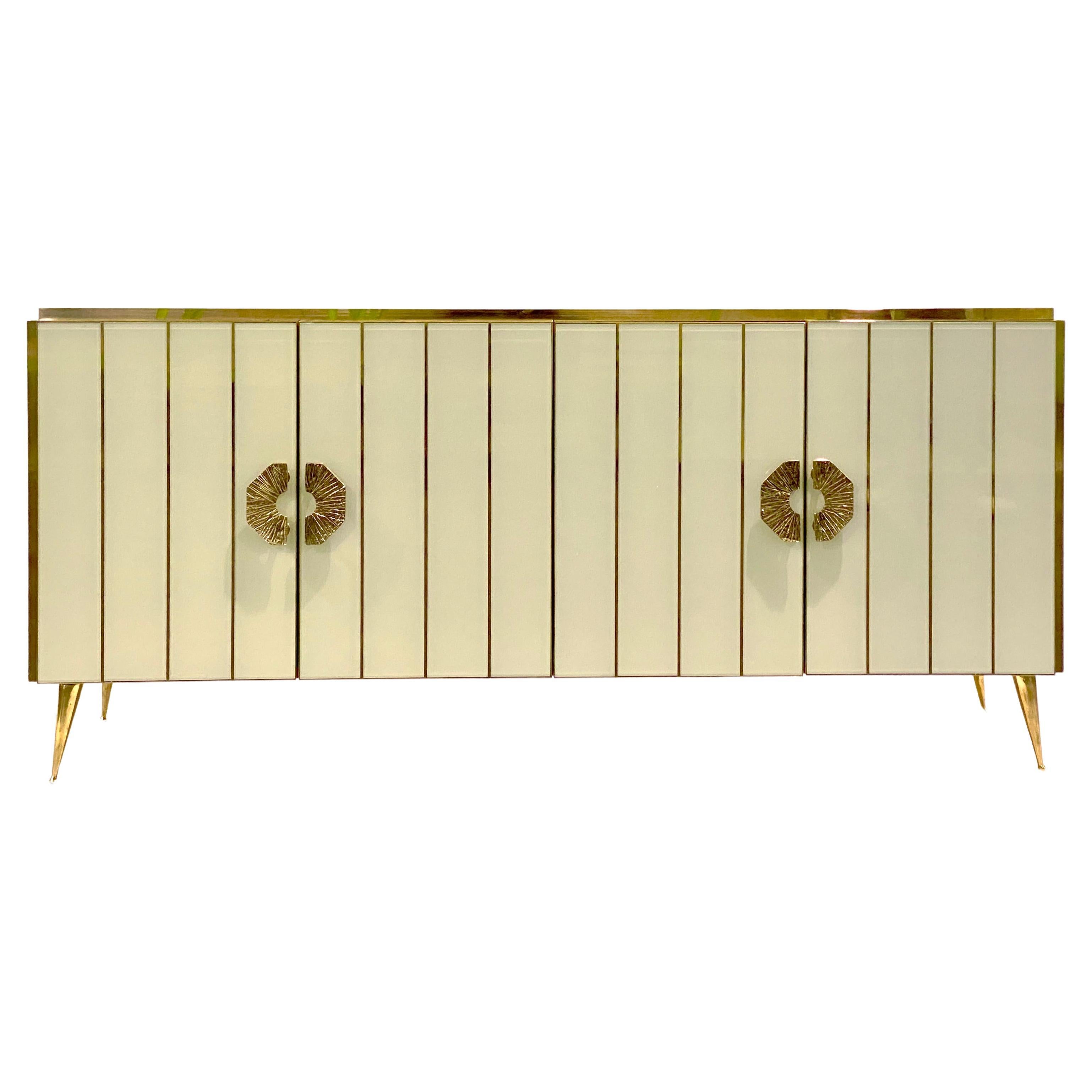 Bespoke customizable 4-door console credenza/sideboard entirely handcrafted in Italy with elegant Art Deco style and Hollywood Regency glam, the surround decorated with art glass in an elegant ivory cream, striped with brass inserts, raised on