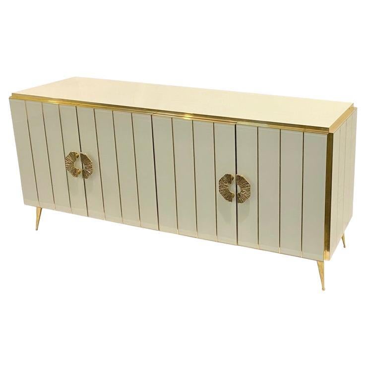 Bespoke customizable 4-door console credenza/sideboard entirely handcrafted in Italy with elegant Art Deco style and Hollywood Regency glam, the surround decorated with art glass in an elegant ivory cream, striped with brass inserts, raised on