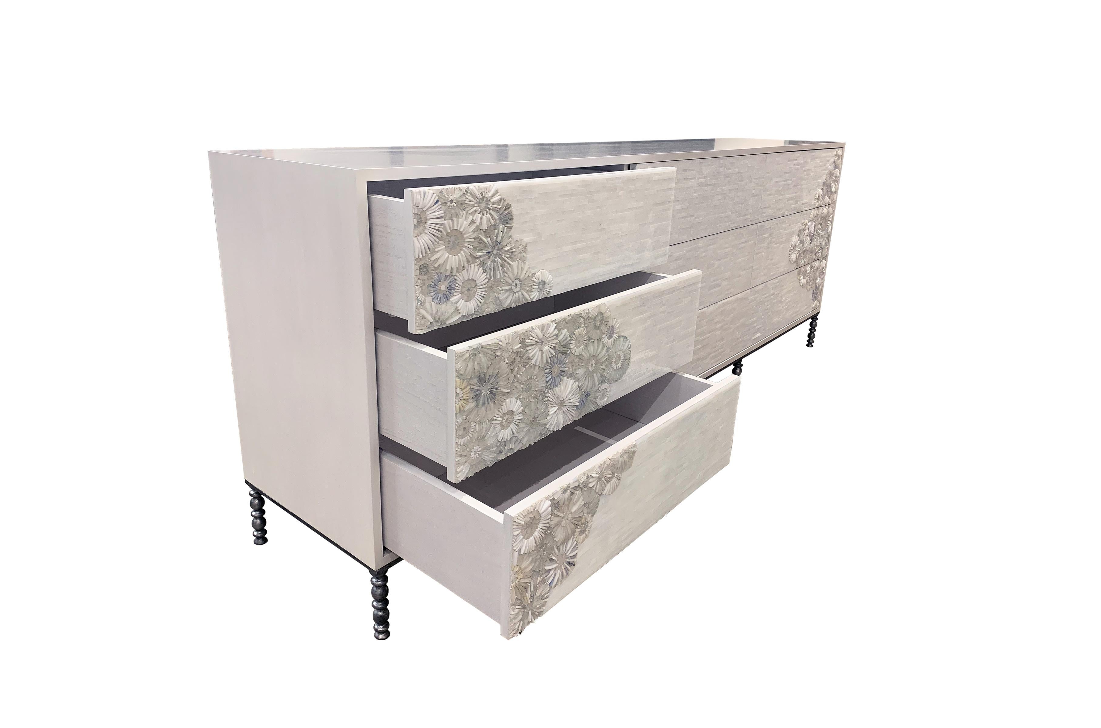 The blossom chest of drawers by Ercole Home has a 9-drawer front, with white wash finish on oakwood.
Handcut glass mosaic in variety shades of white and ivory decorate the surface in blossom and stipe mosaic pattern.
The decorative metal base is