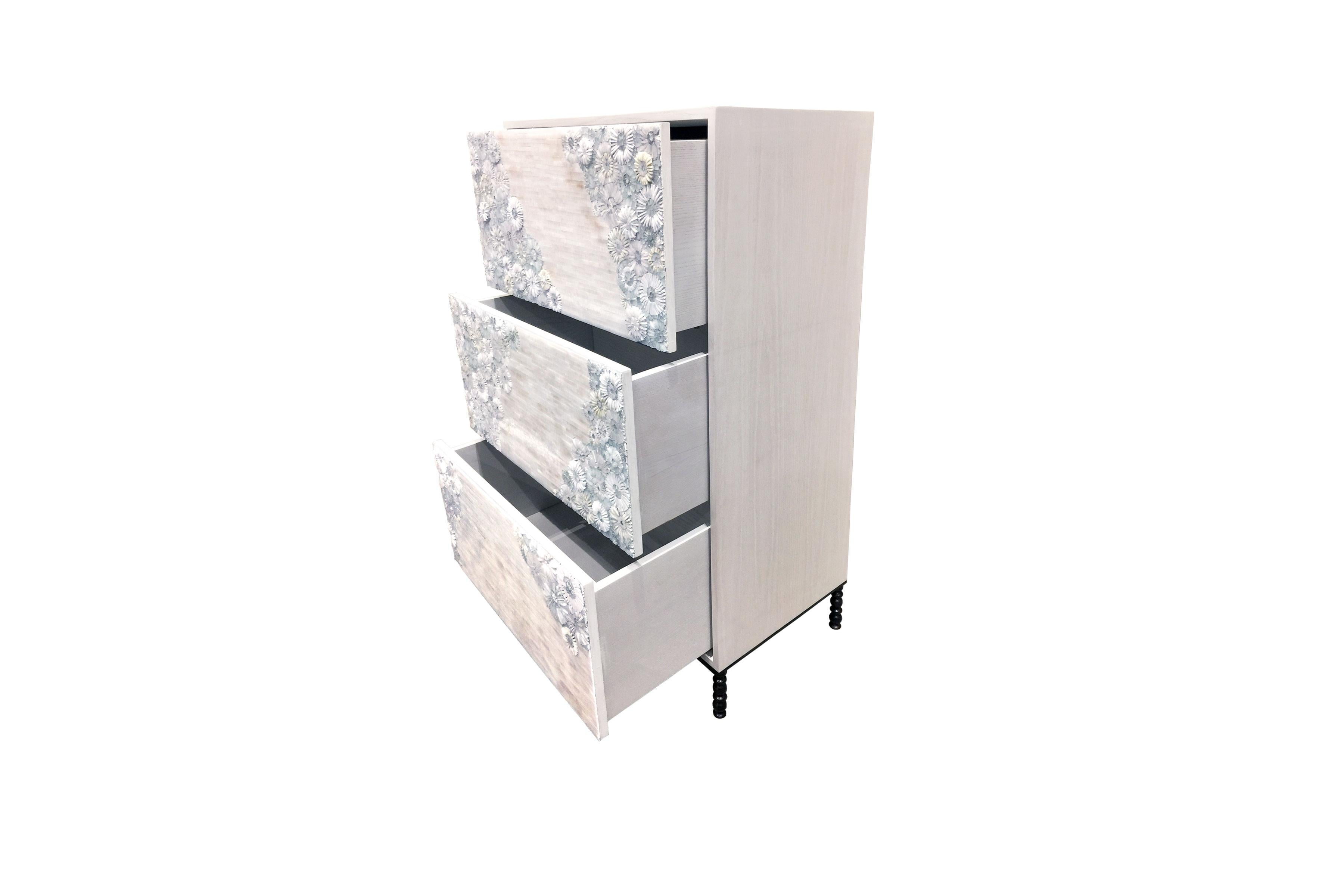 The blossom chest of drawers by Ercole Home has a 3-drawer front, with washed white wood finish on oak.
Handcut glass mosaic in variety shades of white and ivory decorate the surface in blossom and stipe mosaic patterns.
The decorative metal base