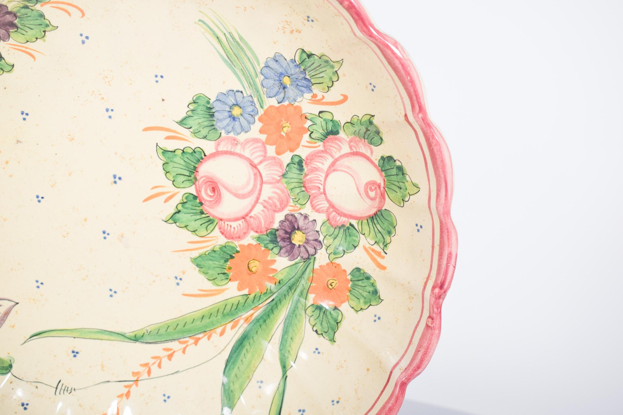 Soucoupe en céramique découpée. Décor polychrome de branches fleuries, bordure festonnée et frise rose.
Le centre de la coupe est décoré d'une belle composition florale, composée de roses dans des tons roses doux, de fleurs orange et violettes, et