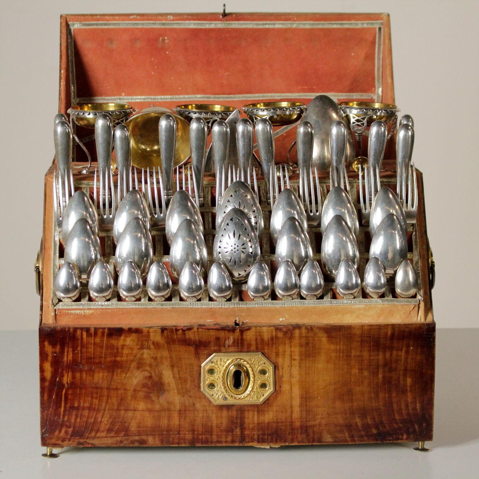 Cherry veneer travel box with brass feet, embossed and gilded metal handles and key hole with neoclassical decorations; the box is covered in red parchment inside and embellished with silver thread trimmings.
The twelve person cutlery set consists