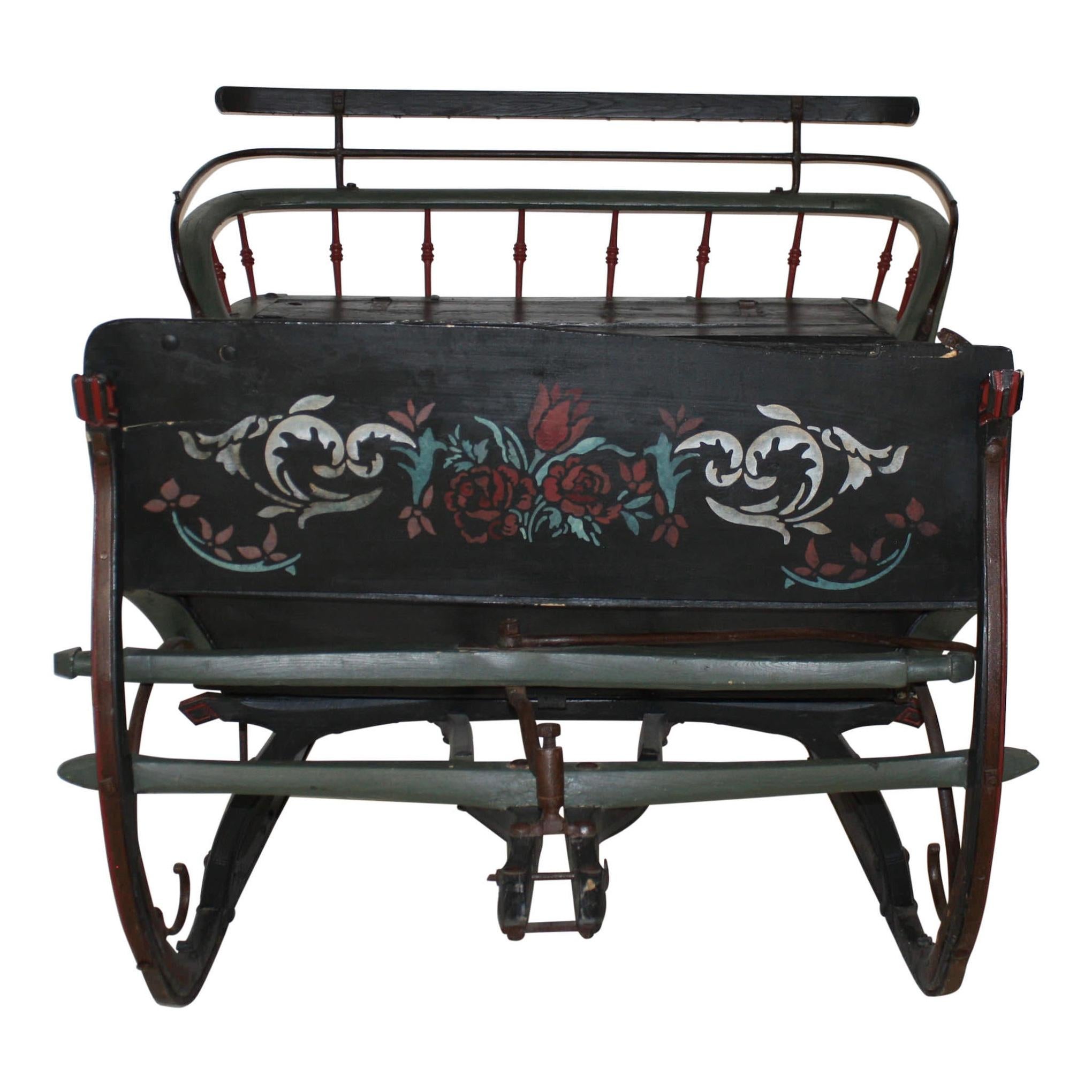 This wooden sleigh features black base paint, green and red trim, and a floral design on the dash. Spindles along the back and scrolled metalwork beneath the seat and floor boards give the sleigh a look that is less weighty than most cutters. The