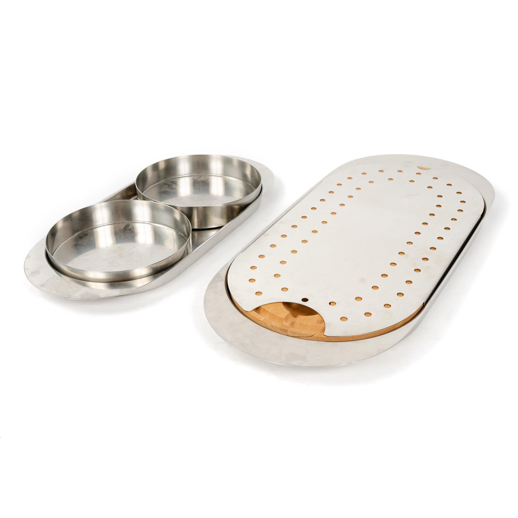 A multifunctional minimalist oval serving and cutting board in polished stainless steel having an inset wooden board and a removable perforated steel cover.