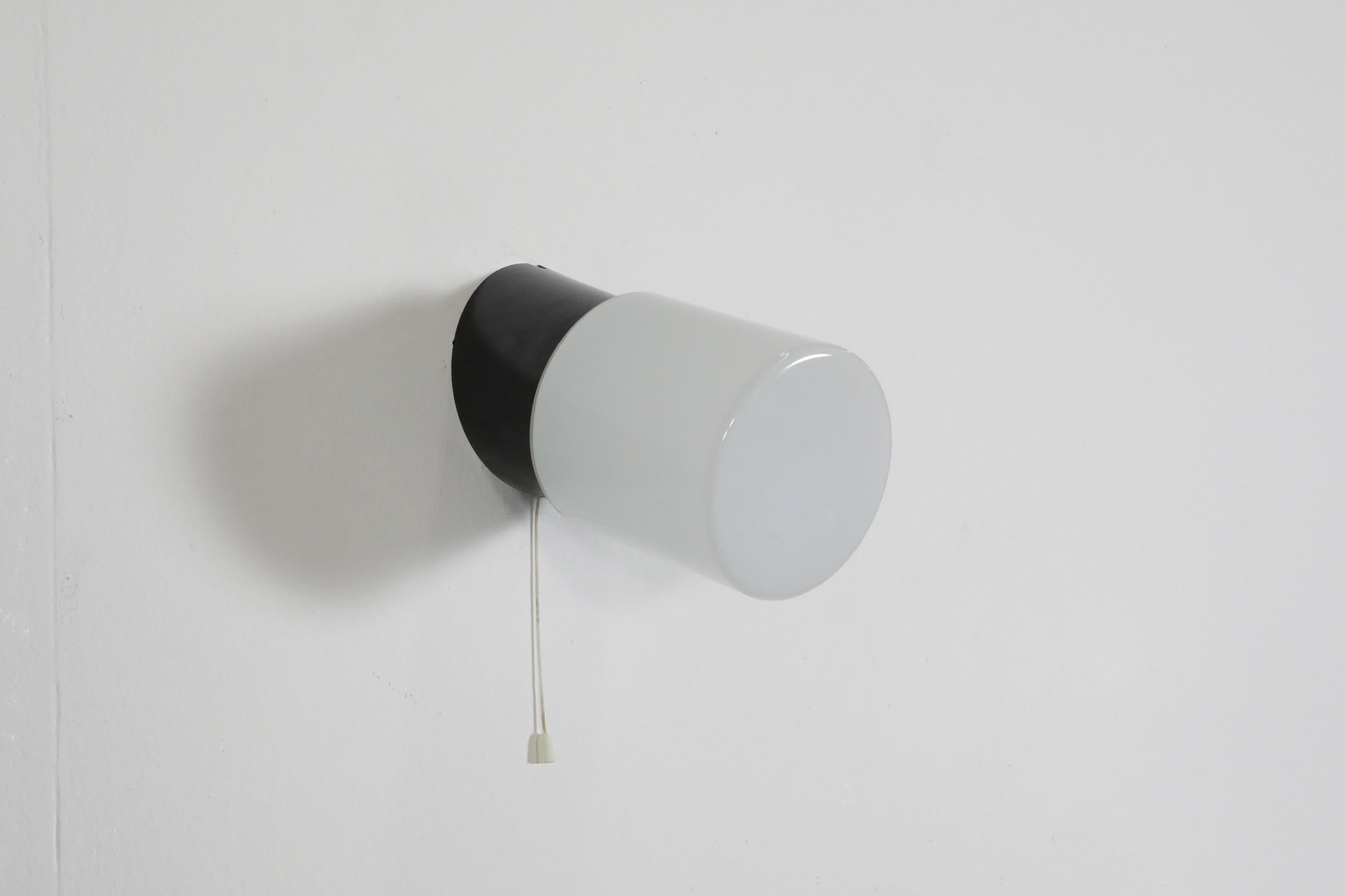 Applique murale cylindrique noire avec base angulaire en bakélite, abat-jour en verre au lait et interrupteur à tirette. Cette lampe est destinée à une installation câblée, mais un cordon peut être ajouté, car il y a une encoche dans la base. En