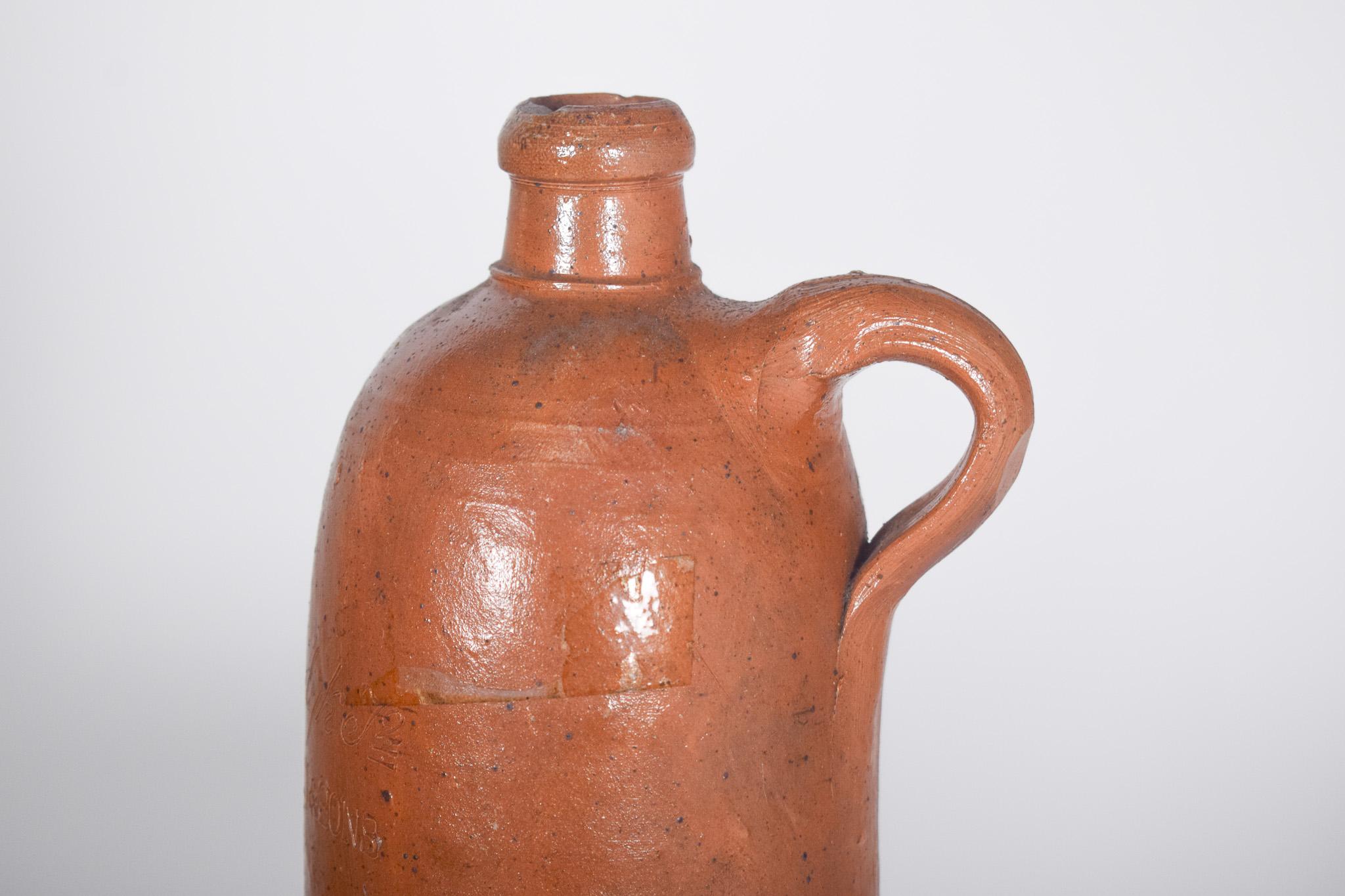 Bottiglia con manico in argilla. Forma cilindrica allungata, con un piccolo collo e un manico laterale. Il pezzo ha un tono terroso caratteristico delle ceramiche tradizionali, con una finitura rustica e una leggera lucentezza dovuta alla smaltatura