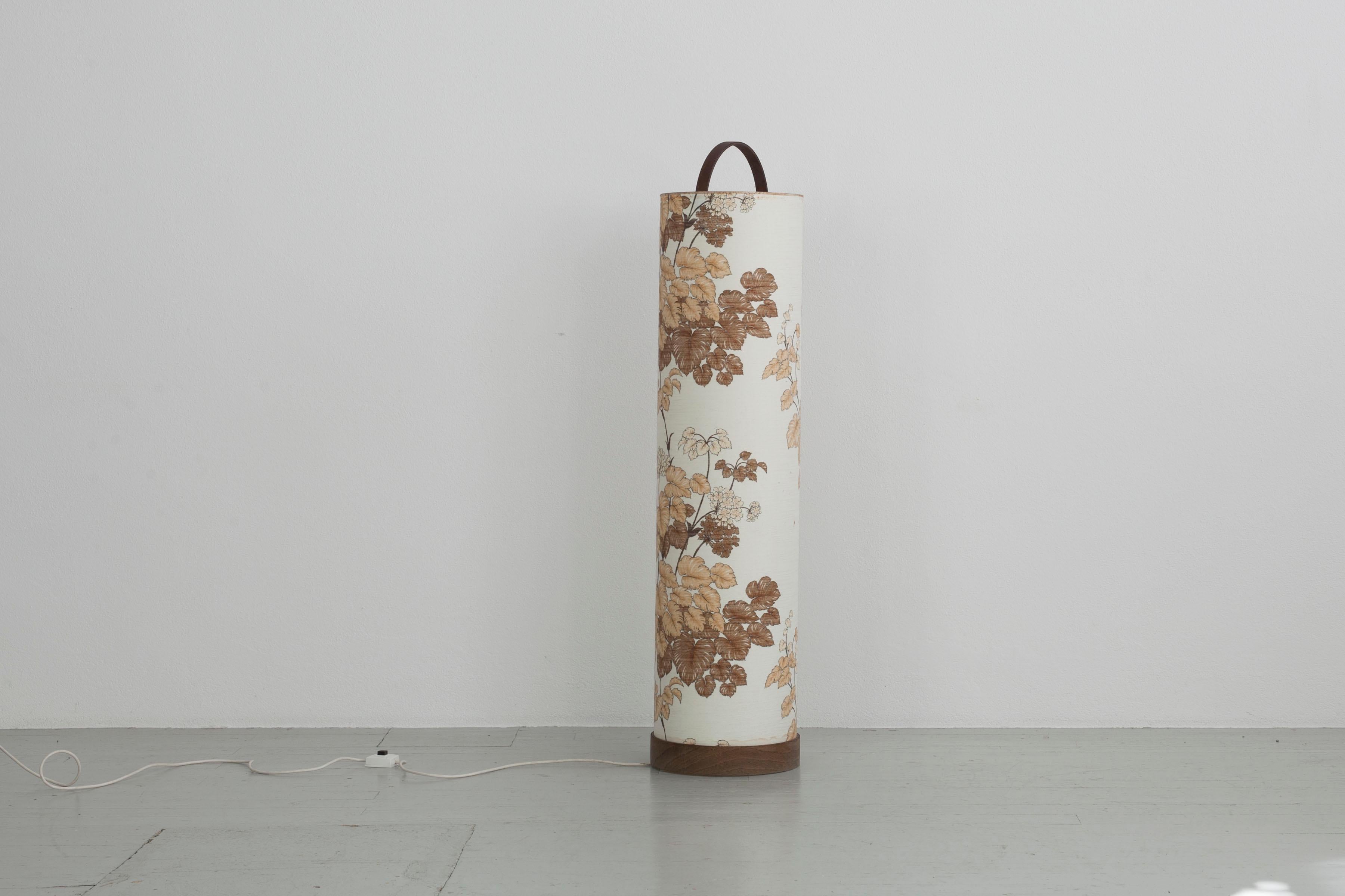 Cylindrical floor lamp from the 70s. The lampshade is continuous to the floor and covered with a floral fabric. The base as well as the handle of the lamp is made of teak wood. It is 132cm high and has a diameter of 31cm. The lamp has an E27 socket.