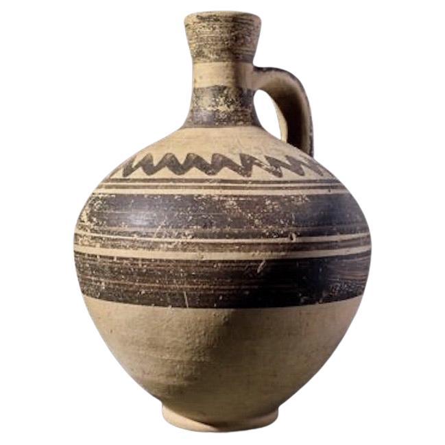 A wheel-thrown pottery jug of ancient Cyprus, formed by an ovoid-biconical body on a low ring foot. The strap handle is arching from the shoulder to the mid-section of a funnel-shaped neck. 
The exterior is decorated with brown painted bands around