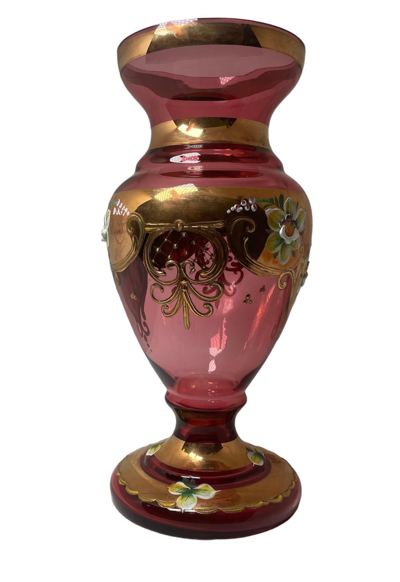 This is a Czech Bohemian Gilt Glass Vase. It depicts a light cranberry color glass vase decorated with hand painted enamel large flowers and gilt scrolls. A gilt scalloped garland with enamel leaves adorned the round base.