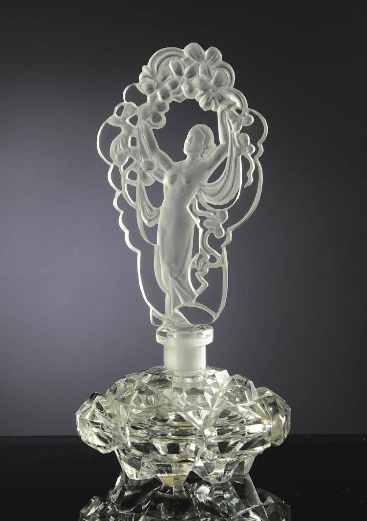 Flacon de parfum et bouchon en verre tchèque transparent avec un autocollant Irice sur le fond du flacon. Le bouchon est en verre percé et décoré d'un nu en verre dépoli entouré de fleurs. Les dimensions sont : 8 1/8