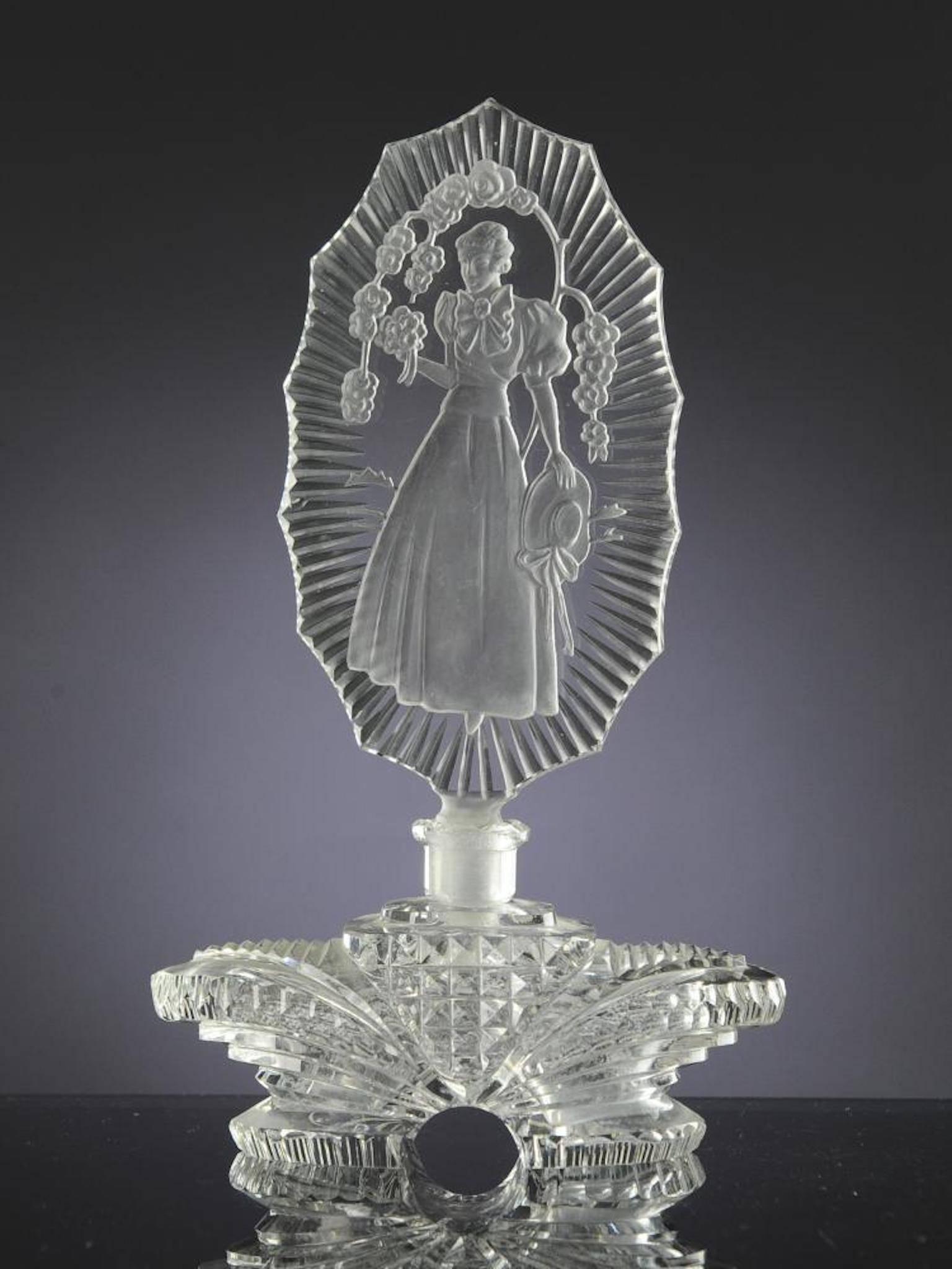 Clear glass perfume bottle and stopper stamped 'Czechoslovakia'. One side of the stopper is decorated with an intaglio woman holding her hat, under a garland of flowers. The other side of the stopper is decorated with cut radiating rays. Dimensions