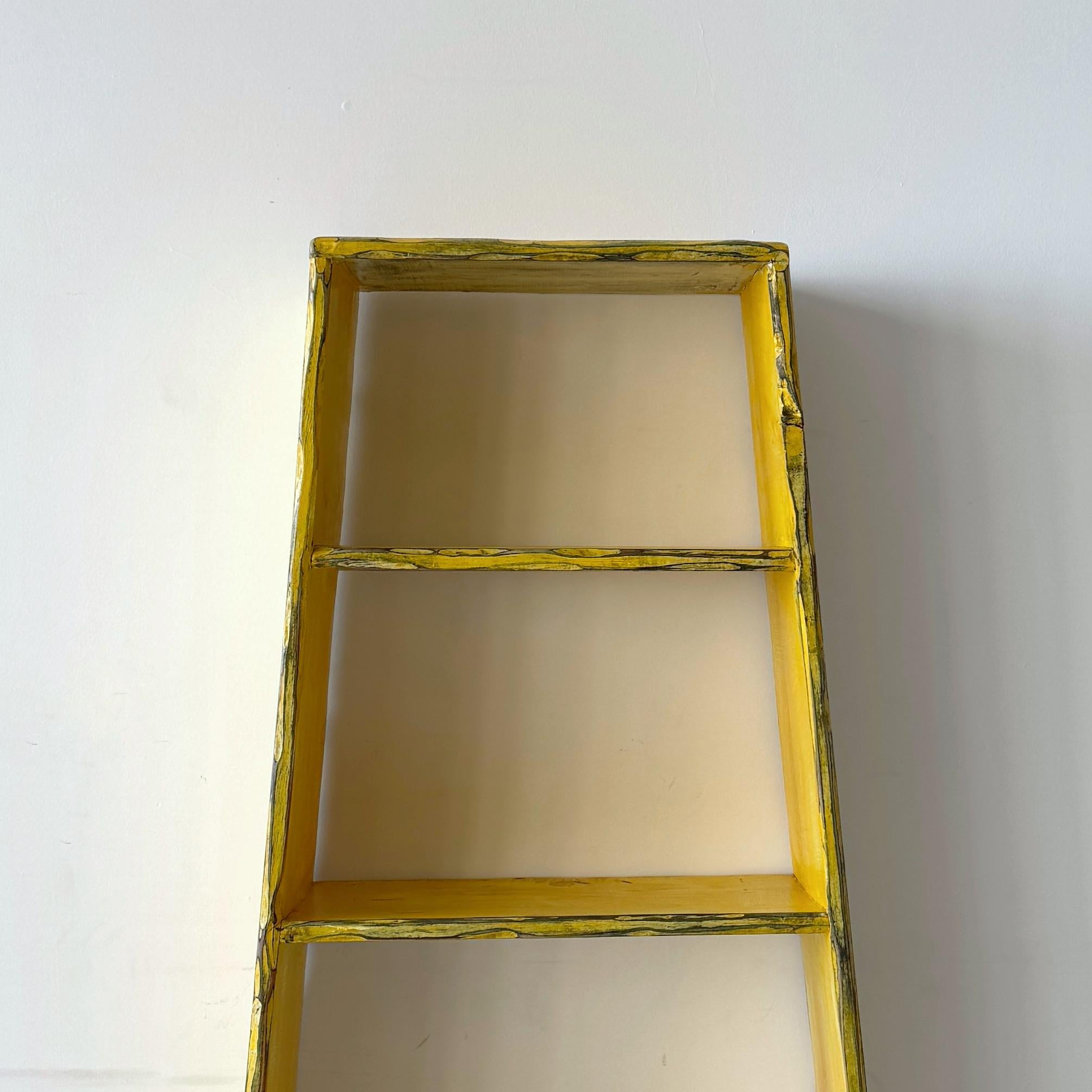 Dreamlike postmodern bookshelf hand-painted in daffodil yellow. The piece features raw, textured edges and sculptural arched cutouts at the base, giving it a handmade, almost architectural presence. Its tapered sides and bright finish make it as
