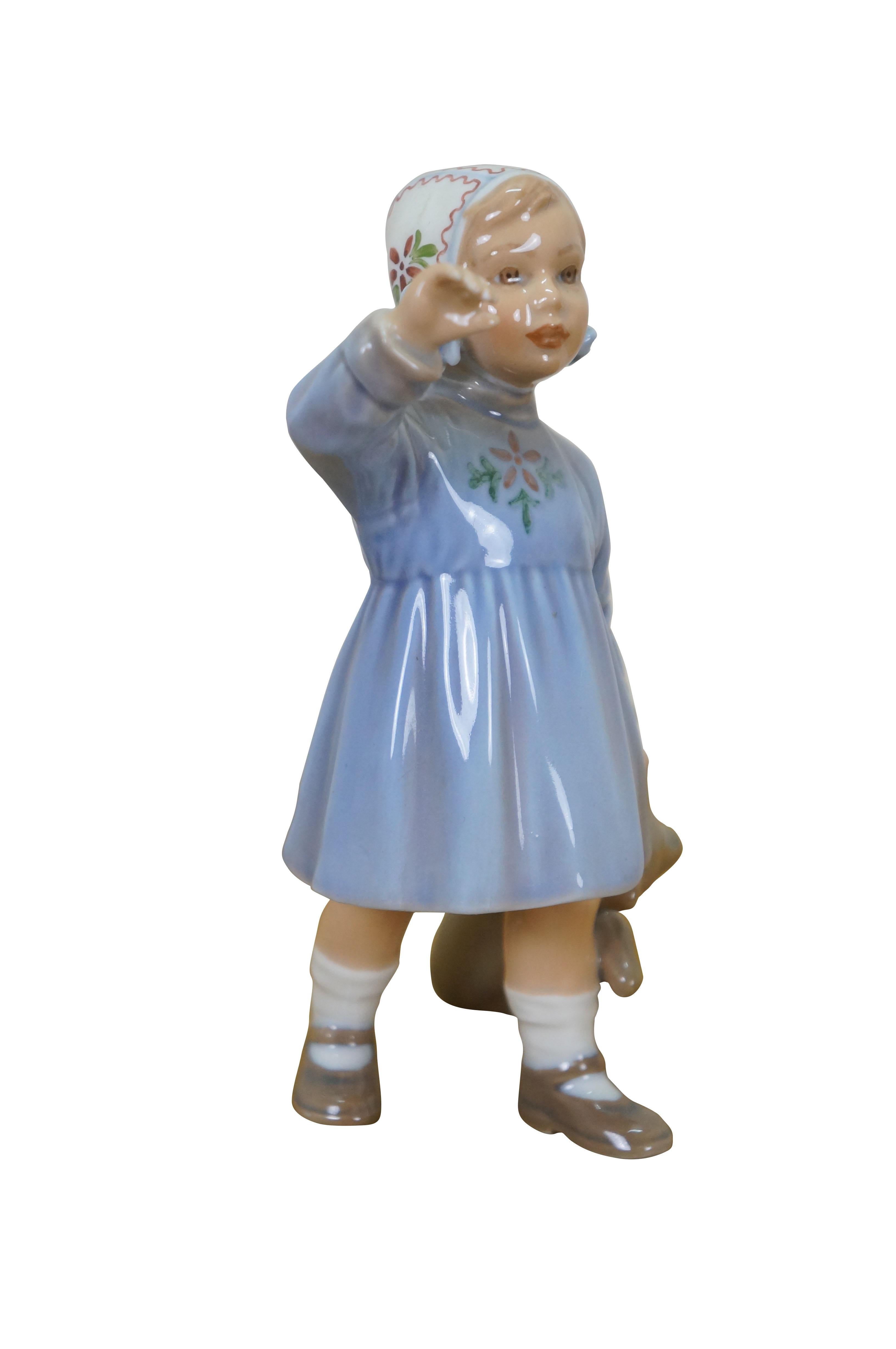 Vintage 20th century porcelain figurine number 1152 by Jens Peter Dahl-Jensen, depicting a young Danish girl in a blue dress and floral bonnet, holding a teddy bear in one hand and raising the other in the air.

Dimensions:
3