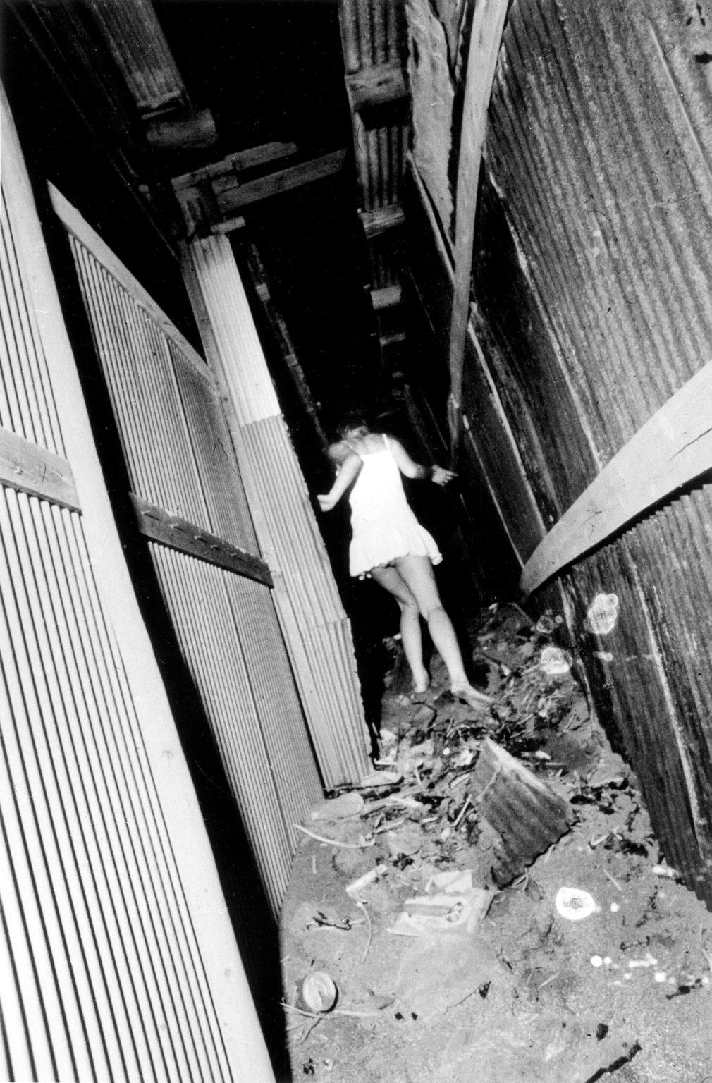 Photographie signée Daido Moriyama : Yokosuka, une ville japonaise 1971/2020 :

Une représentation granuleuse, en noir et blanc et très contrastée, signée Daido Moriyama, d'une jeune fille anonyme en robe blanche courant dans une ruelle pleine de