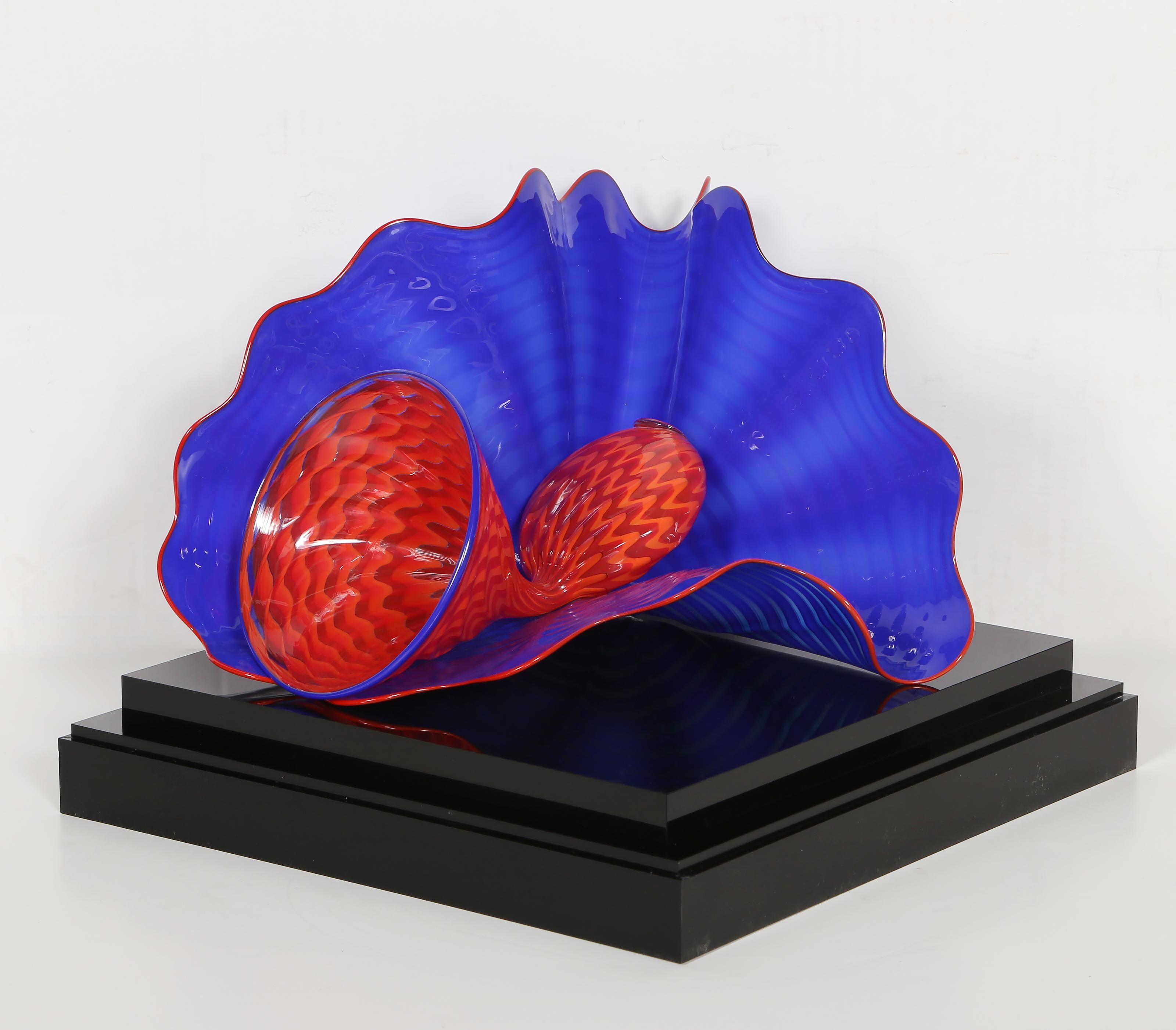 En esta magnífica escultura soplada a mano por el artista estadounidense Dale Chihuly, un vibrante objeto rojo se acuna suavemente en el hueco curvado de una ondulante concha azul. Esta pieza única procede de la serie Persas de Chihuly, una