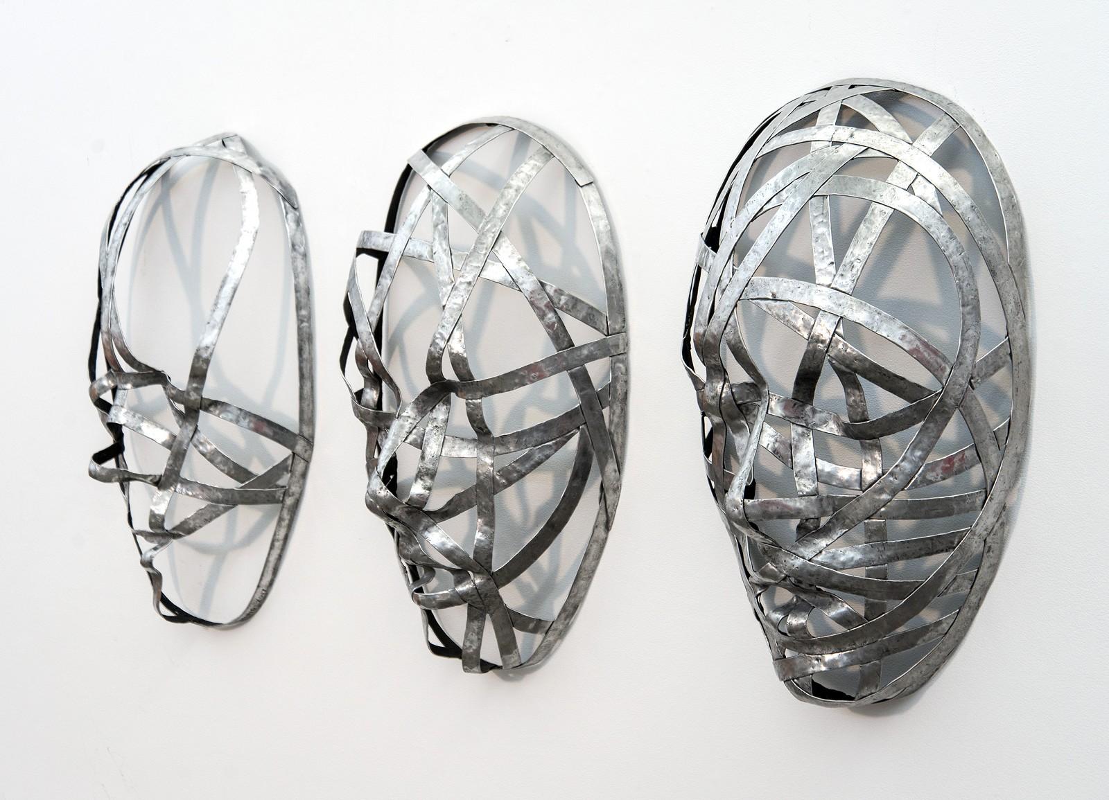 D'étroites bandes d'aluminium sont habilement façonnées pour former les courbes et les plis de trois masques du sculpteur Dale Dunning. Les visages enveloppés d'inspiration industrielle steam-punk qui changent de droite à gauche reflètent également