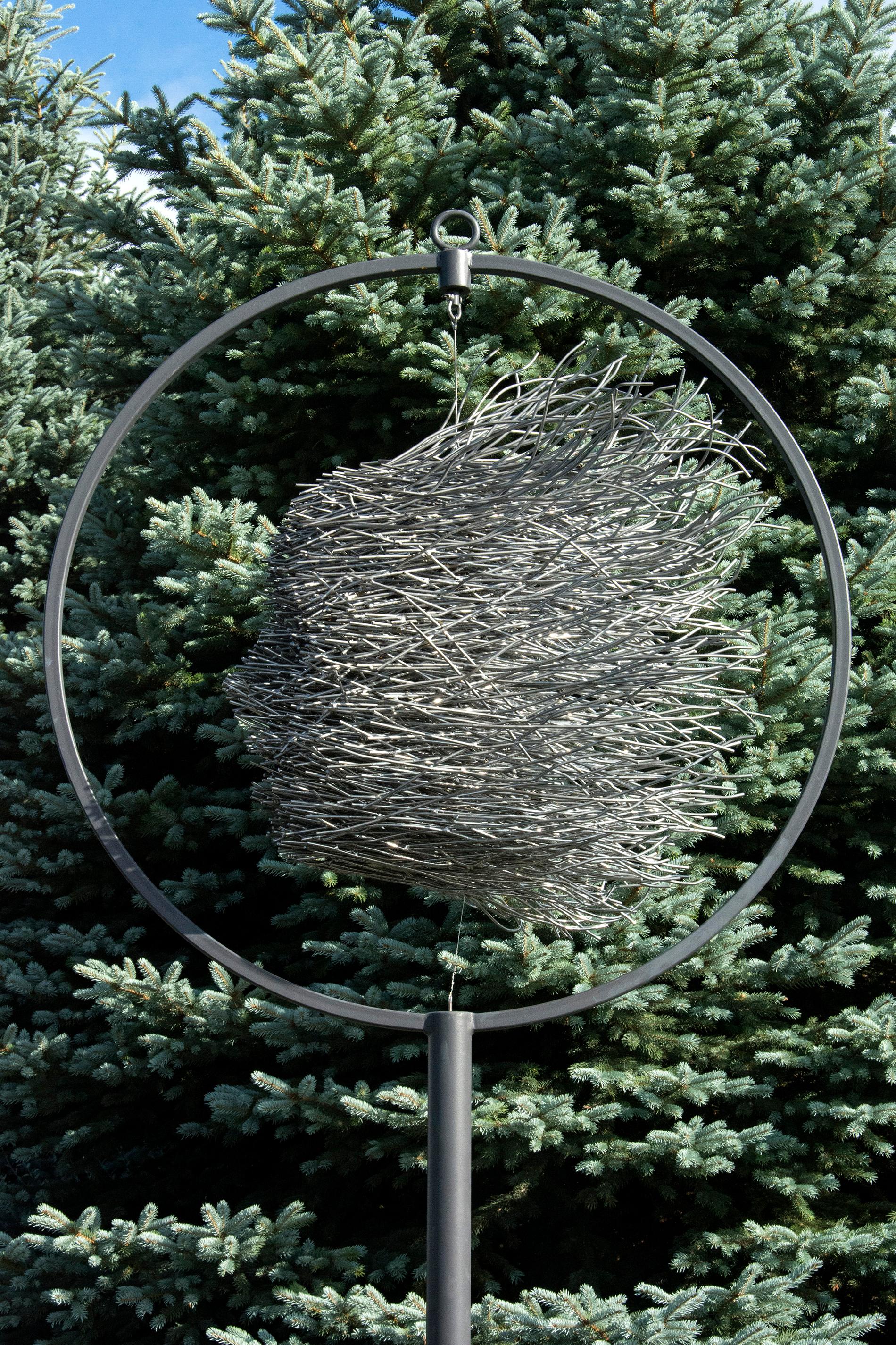 Escultura cinética de exterior. Un rostro silencioso emerge de una maraña de brillantes varillas de acero con recubrimiento de polvo en esta escultura de exterior de Dale Dunning. La cara está suspendida en el centro de un anillo perfecto encaramado