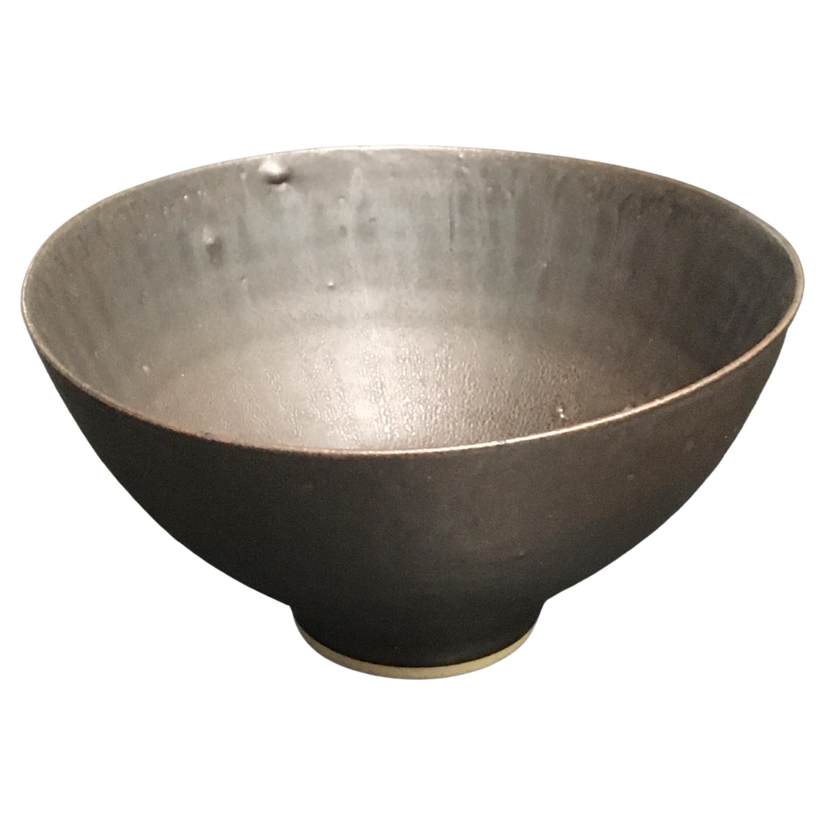 This is a great example of a Lucie Rie iconic hand-thrown, footed bowl with manganese glaze.

It is thinly potted and slightly pulled into an ovoid shape with an undulating rim.

The running glaze is subtle whilst adding an individual element to the