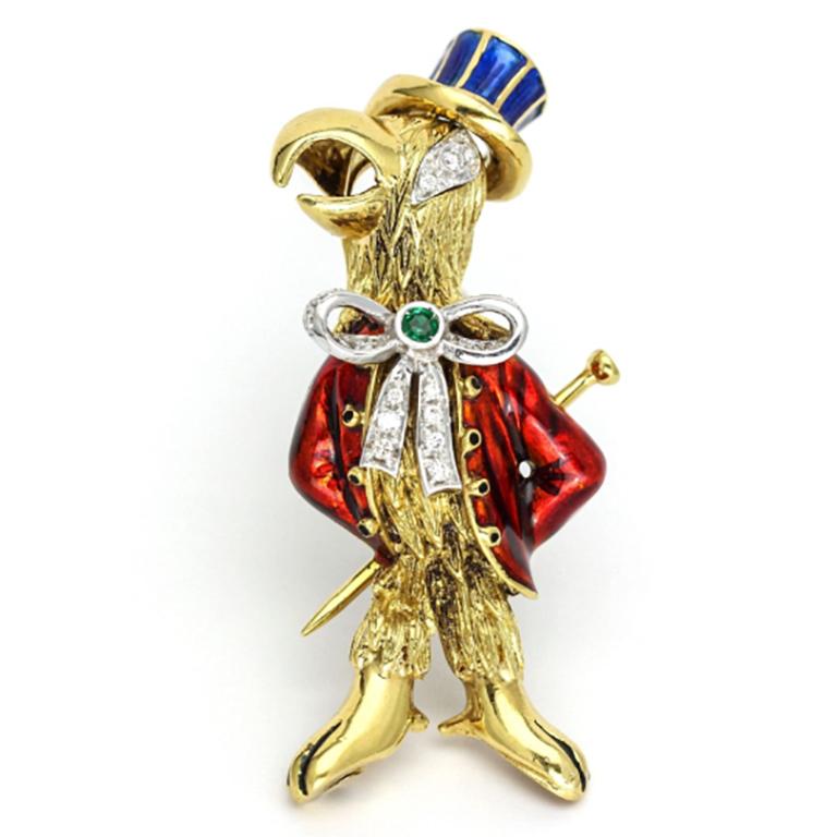 Damiani Parrot brooch with red and blue enamel details on clothes and hat. The bird is wearing a bow tie set with pave diamonds and a bezel set emerald detail at the center. The eye is encrusted with diamonds. The brooch comes with a pyrite stand