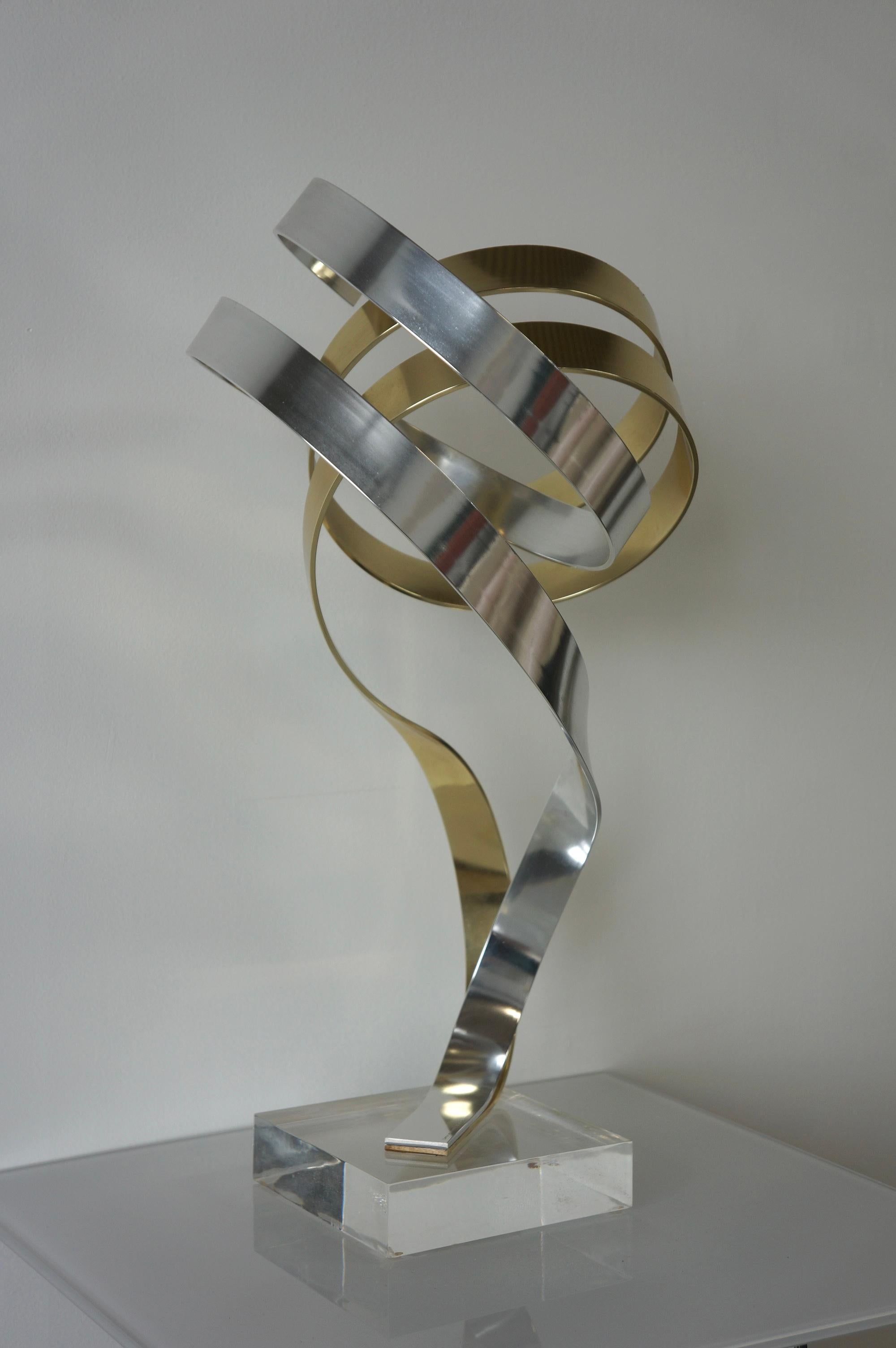 Abstract ‘Ribbon’ sculpture made with anodized aluminum with gold tone by Dan Murphy. This kinetic sculpture is reminiscent of a ribbon shape in which the aluminum sculpted pieces are intertwined supported by a lucite base. It’s dated and signed Dan