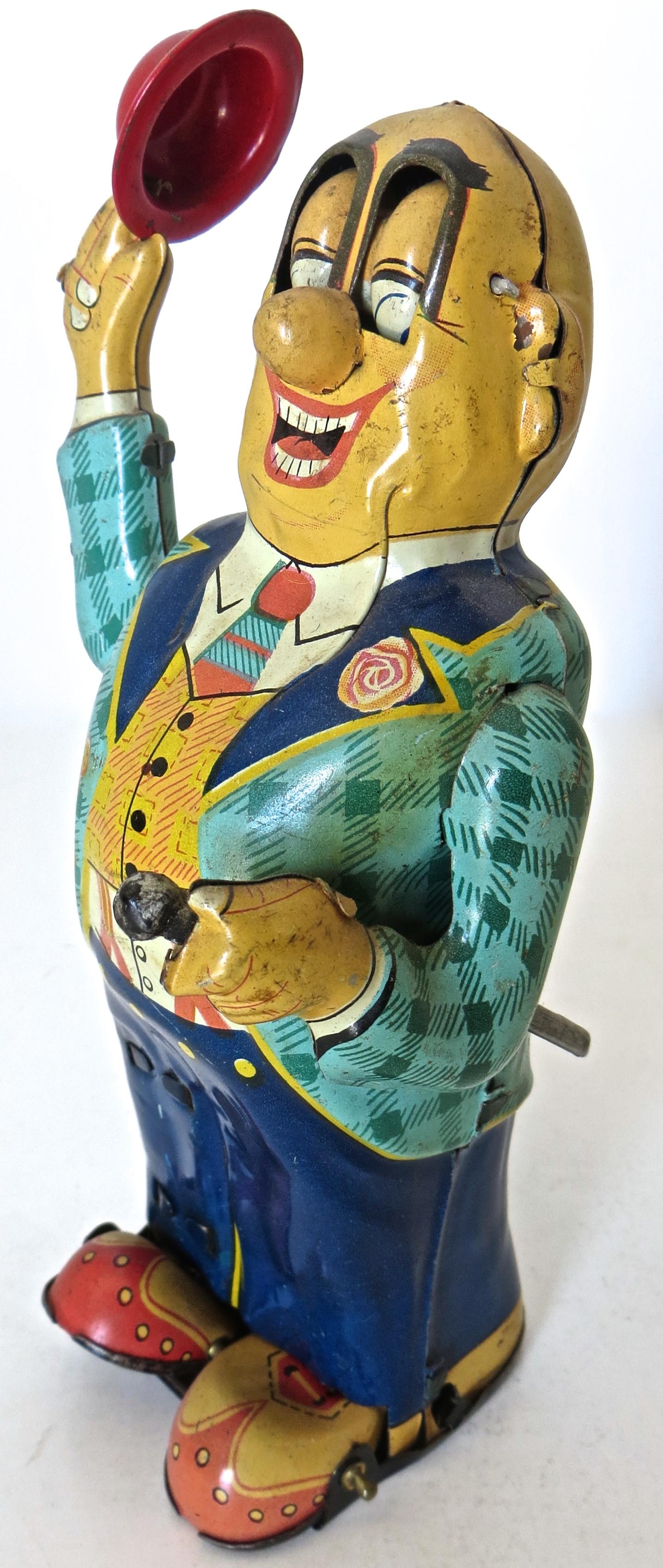 This wonderful, charming and pleasing action wind up toy was manufactured by HTC Mikuni Company of Japan, circa 1950s. It is made completely of tin, and is colorfully lithographed in green, blue, white, and flesh tones, with a tin red hat. The